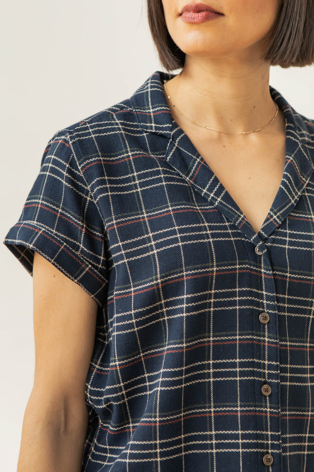 A woman wears the Bridge & Burn Innes Shirt in Dusk Herringbone Plaid—a relaxed fit, navy blue plaid short-sleeve button-up with red and white lines—paired with a simple gold necklace. Her chin-length bob frames her visible torso and lower face.