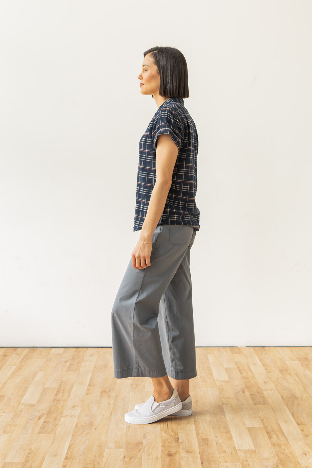 A woman with short dark hair stands in profile on a wood floor wearing the Bridge & Burn Innes Shirt in Dusk Herringbone Plaid, paired with gray wide-leg pants and white slip-on shoes, against a plain white background.