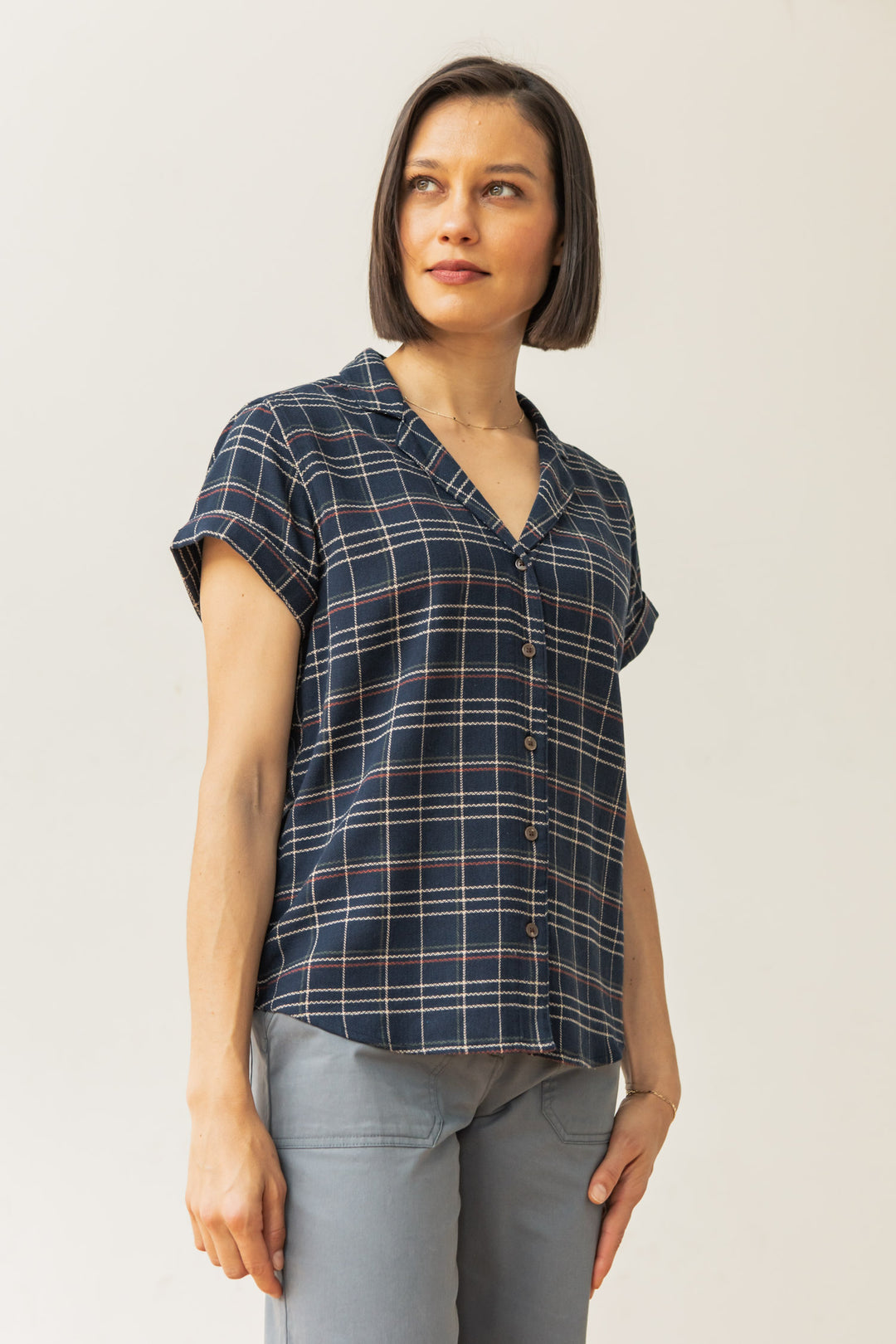 A woman with a bob wears the Bridge & Burn Innes Shirt in Dusk Herringbone Plaid, a relaxed-fit recycled cotton short-sleeve button-up, paired with light gray pants, standing against a plain light background looking slightly left.
