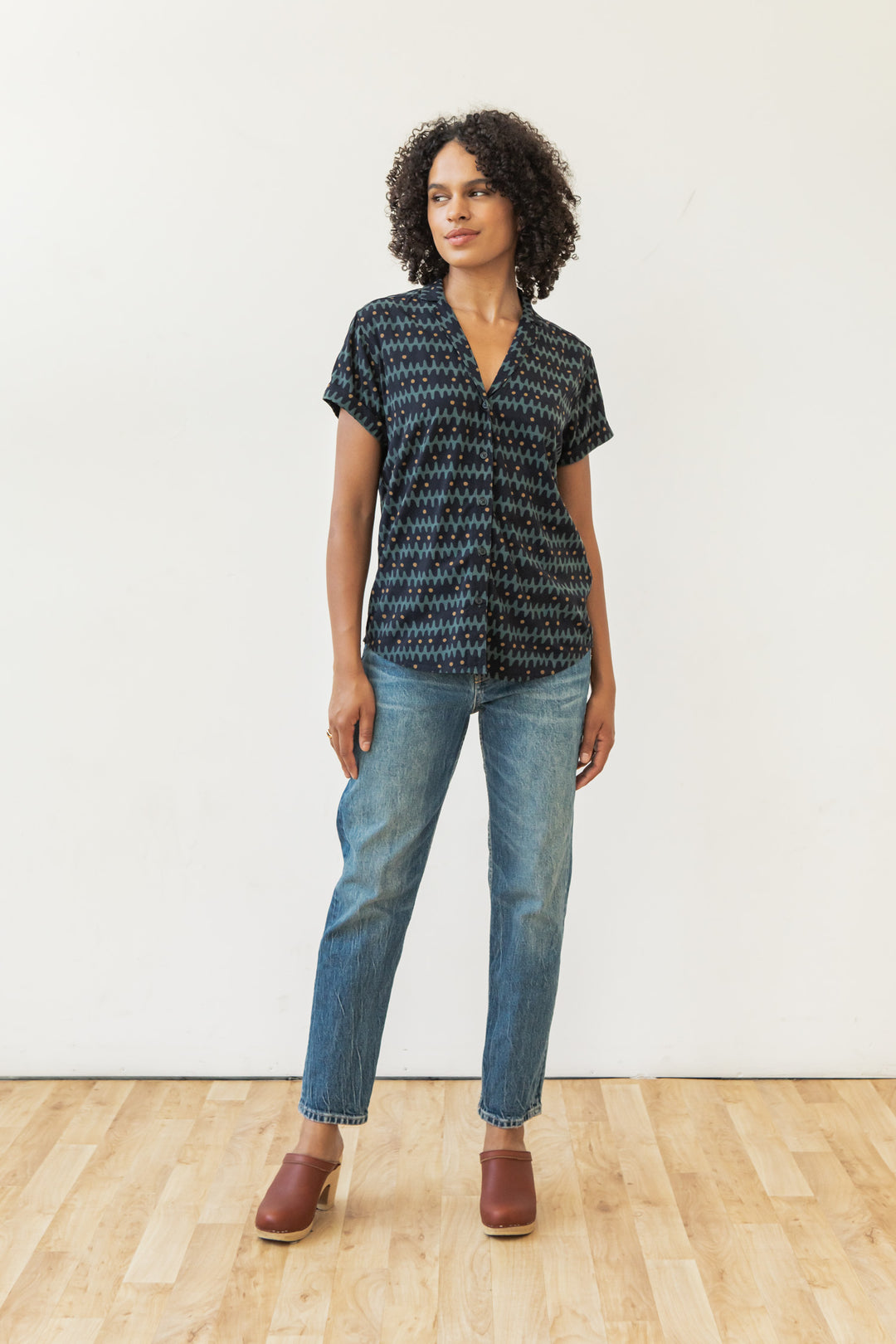 A woman with curly hair stands on a wooden floor against a white wall, wearing the Bridge & Burn Innes Shirt in Fishbone Ink, blue jeans, and brown clogs.