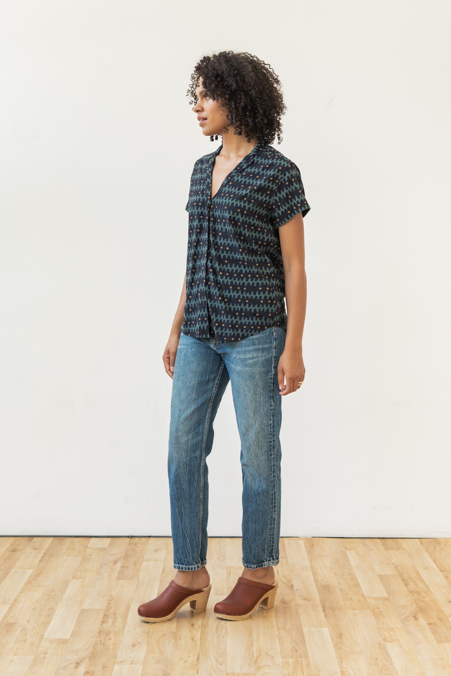 A person with curly hair stands on a wooden floor against a white wall, wearing the Bridge & Burn Innes Shirt in Fishbone Ink, paired with blue jeans and brown clogs.