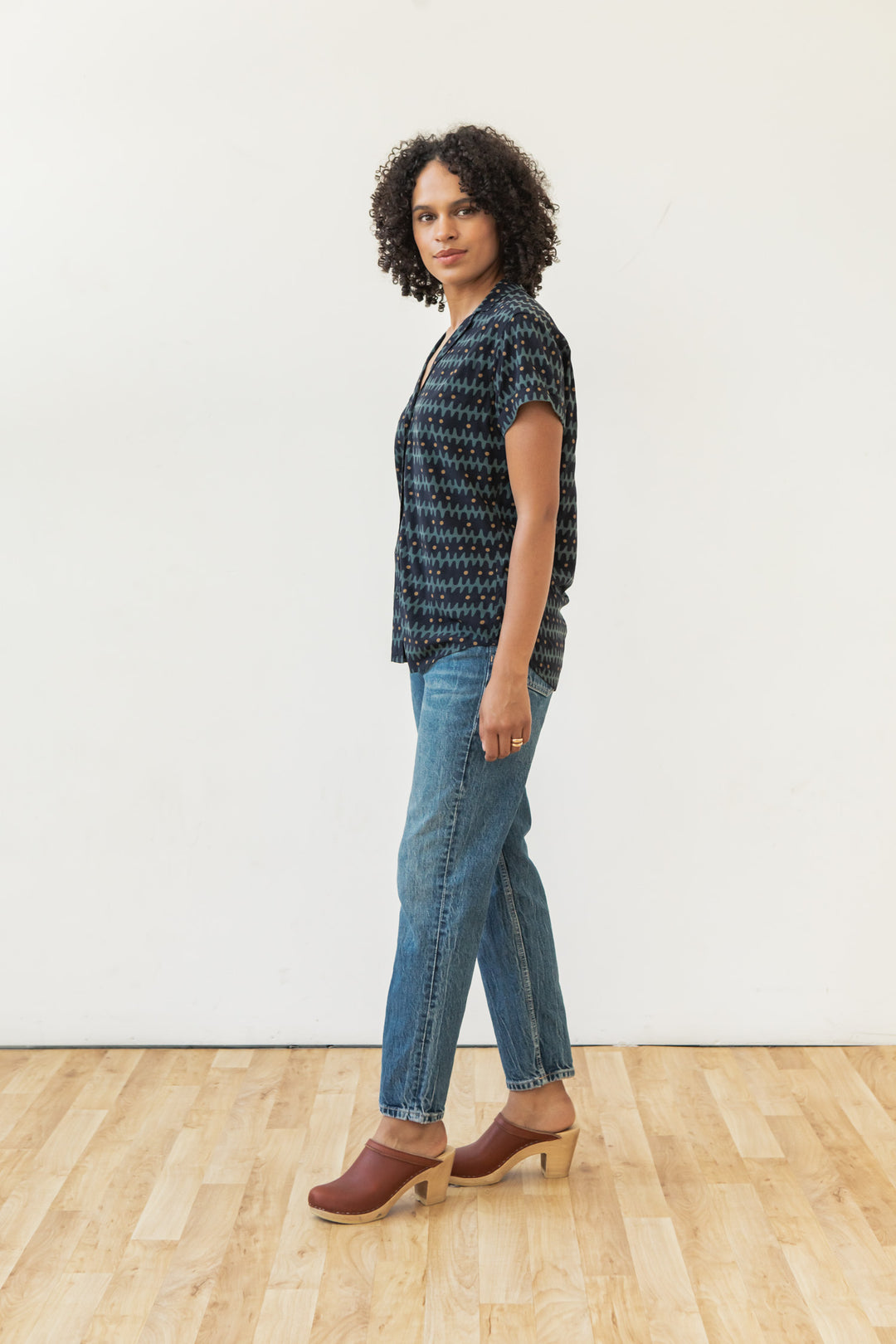 A person with curly hair stands sideways on a wooden floor, wearing the Bridge & Burn Innes Shirt in Fishbone Ink, blue jeans, and brown heeled clogs, posed against a plain white wall.