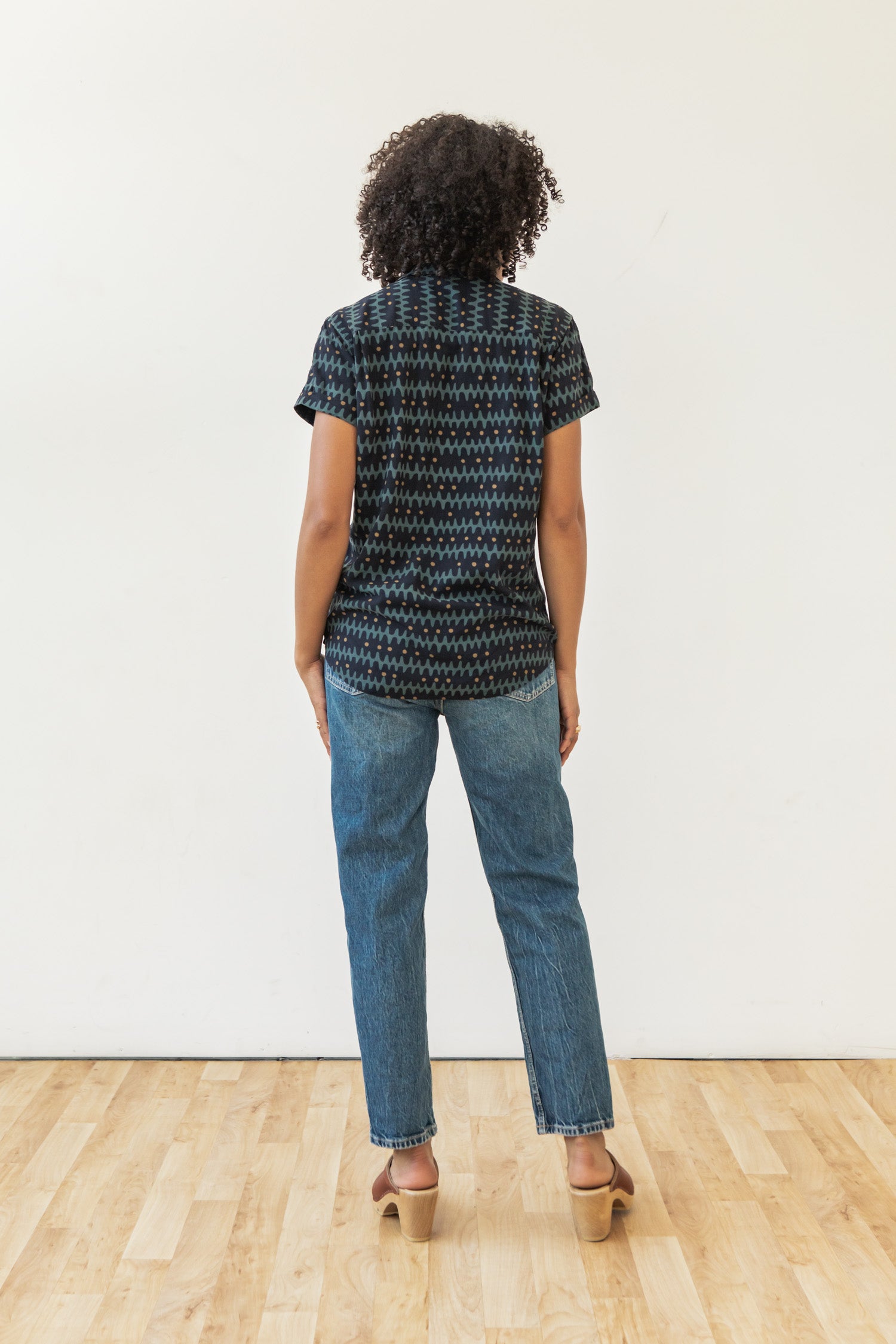 Facing away from the camera, a person with curly hair stands indoors on a wood floor against a white wall, wearing the Bridge & Burn Innes Shirt in Fishbone Ink with blue jeans and tan heeled shoes.