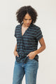 A person with curly hair wears the Bridge & Burn Innes Shirt in Fishbone Ink and blue jeans, looking down with hands in their pockets, standing against a plain light background.