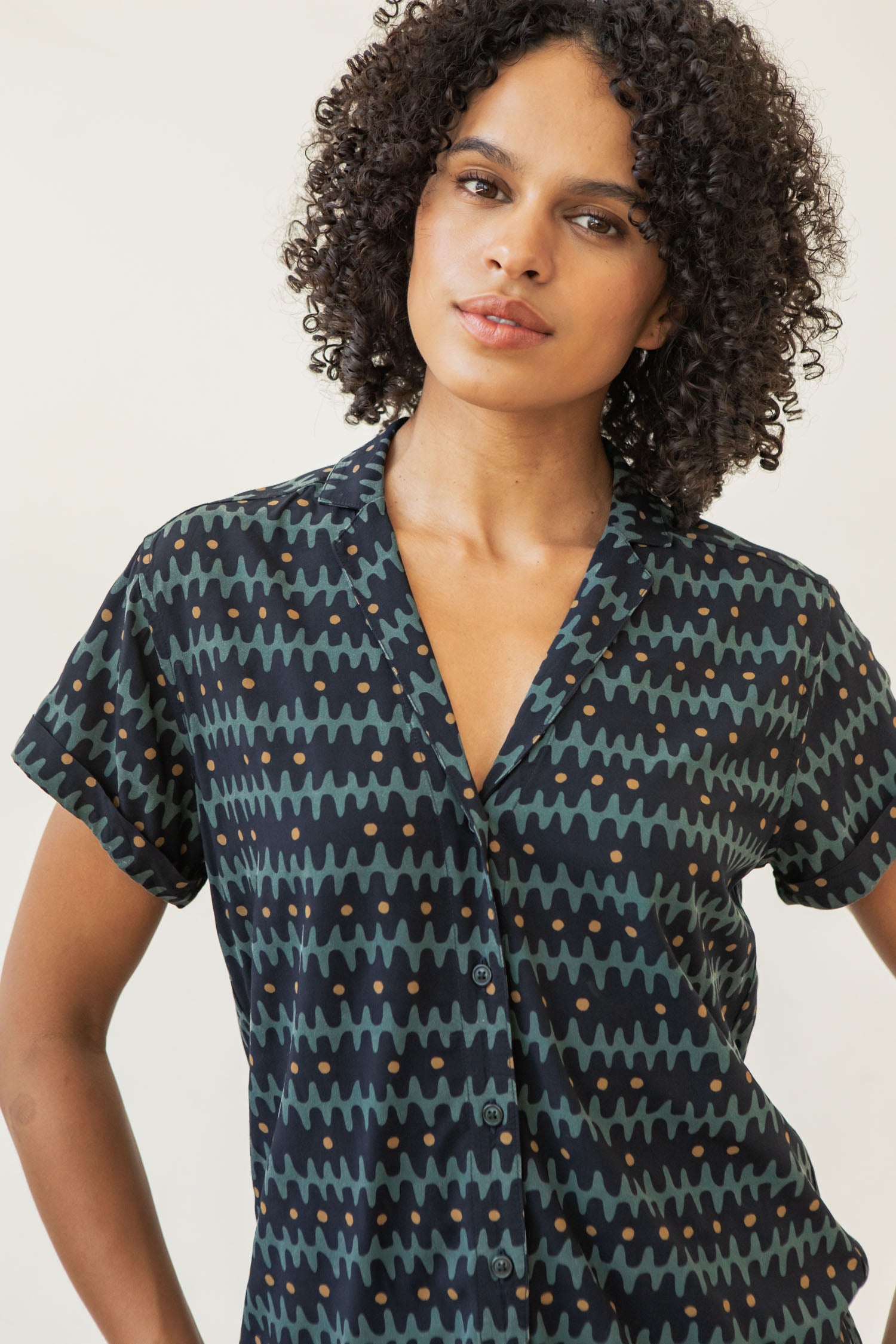 A woman with curly hair wears the Bridge & Burn Innes Shirt in Fishbone Ink, a relaxed fit, short-sleeve button-up with blue, black, and olive green zigzag and dot patterns and a lapel collar, standing against a light background.