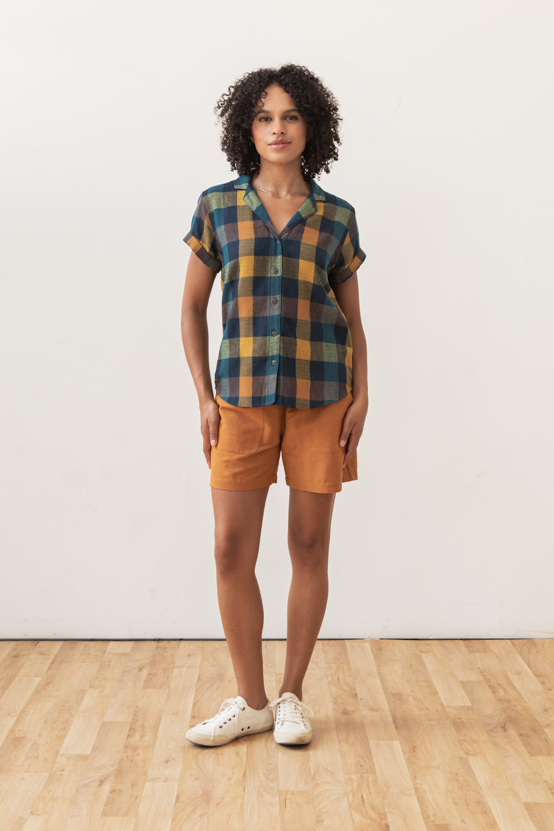A person stands indoors on a wooden floor against a white wall, wearing the Bridge & Burn Innes Shirt in Moonbow Plaid— a relaxed fit button-up made from recycled cotton—paired with orange shorts and white sneakers.