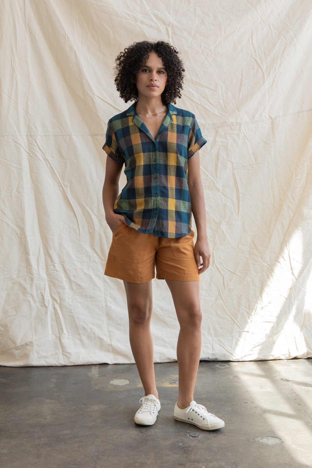 A person with curly hair stands against a cream fabric backdrop, wearing the Bridge & Burn Innes Shirt in Moonbow Plaid, tan shorts, and white sneakers, with one hand in their pocket.