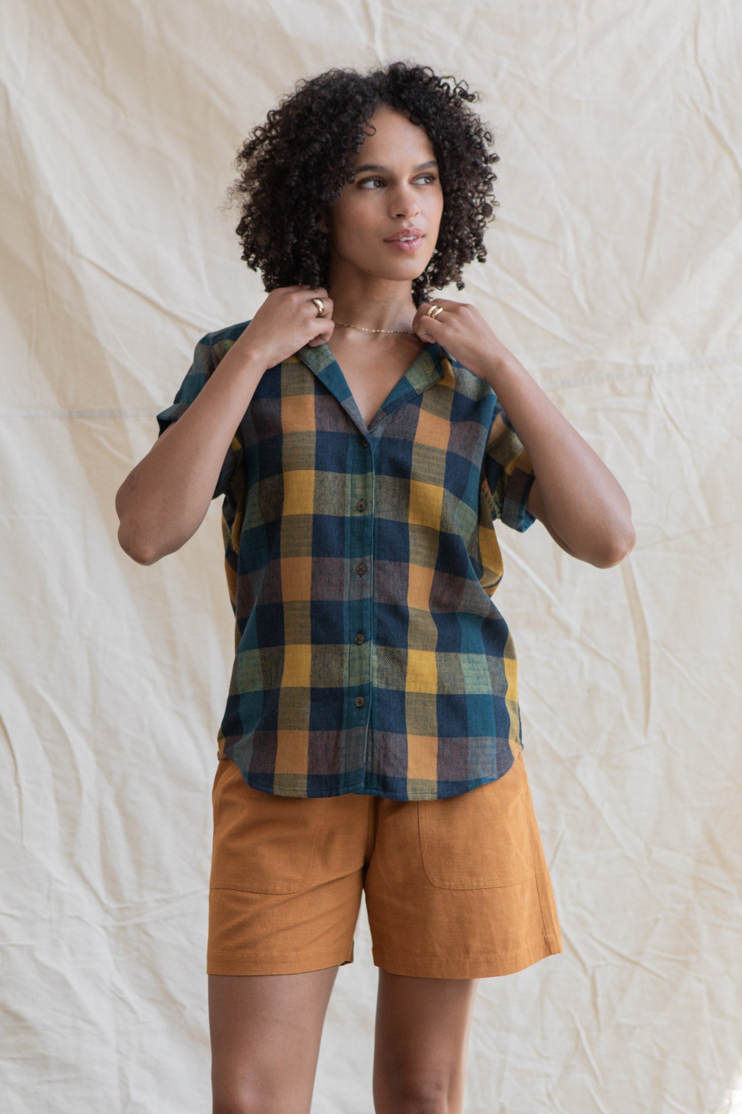 A person with curly hair wears the Bridge & Burn Innes Shirt in Moonbow Plaid, made from recycled cotton, paired with rust-colored shorts, standing before a cream fabric backdrop.