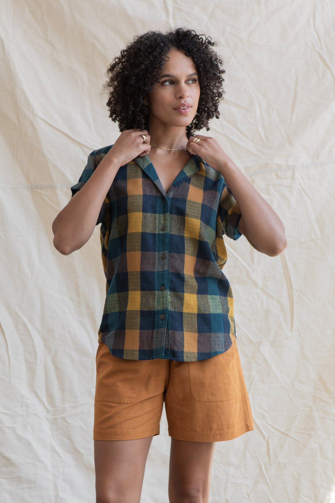 A person with curly hair wears the Bridge & Burn Innes Shirt in Moonbow Plaid, made from recycled cotton, paired with rust-colored shorts, standing before a cream fabric backdrop.