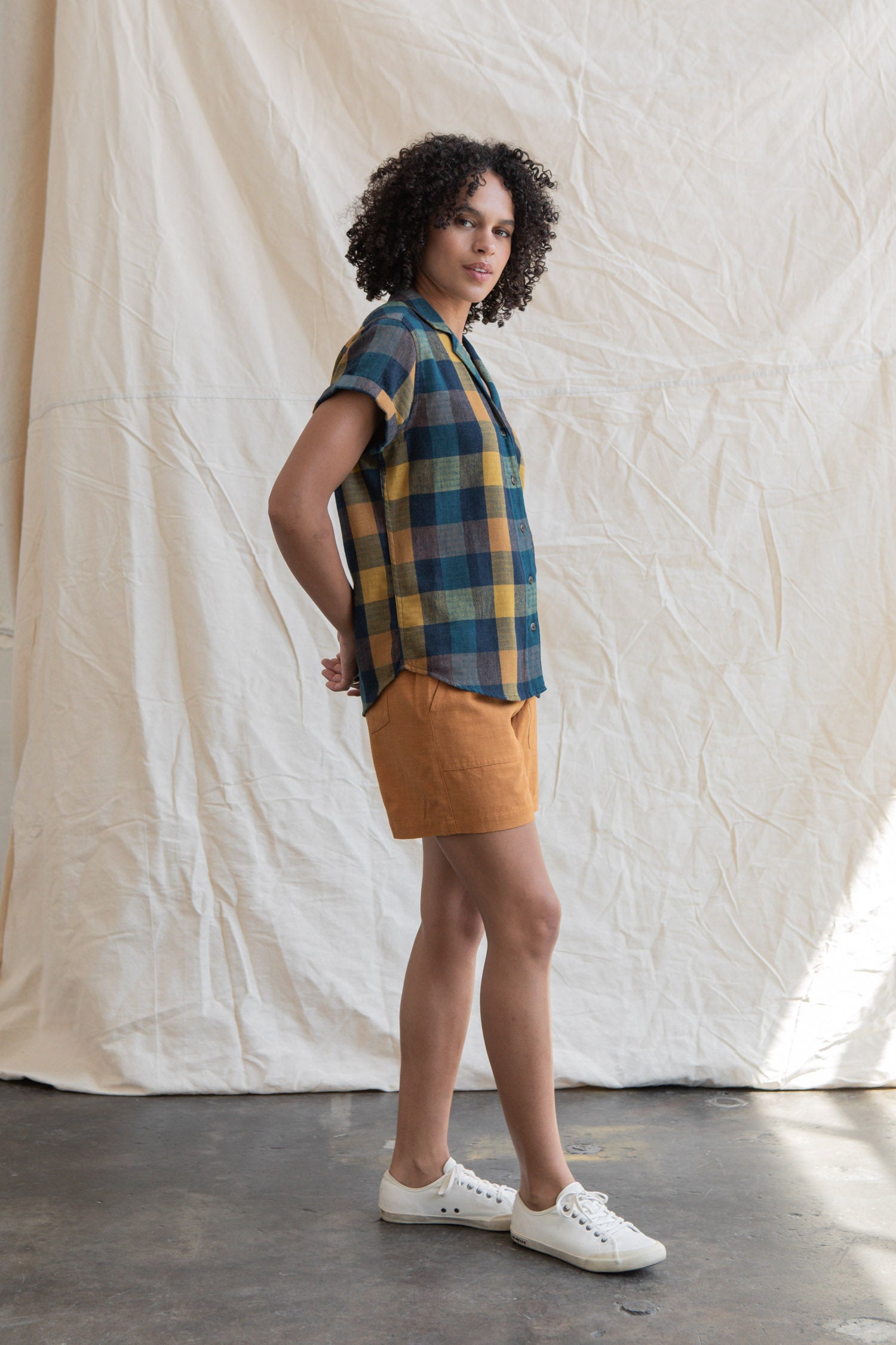A person with curly hair stands sideways before a beige fabric backdrop, wearing the Bridge & Burn Innes Shirt in Moonbow Plaid, orange shorts, and white sneakers.