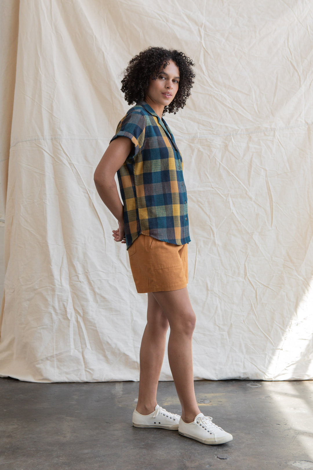 A person with curly hair stands sideways before a beige fabric backdrop, wearing the Bridge & Burn Innes Shirt in Moonbow Plaid, orange shorts, and white sneakers.
