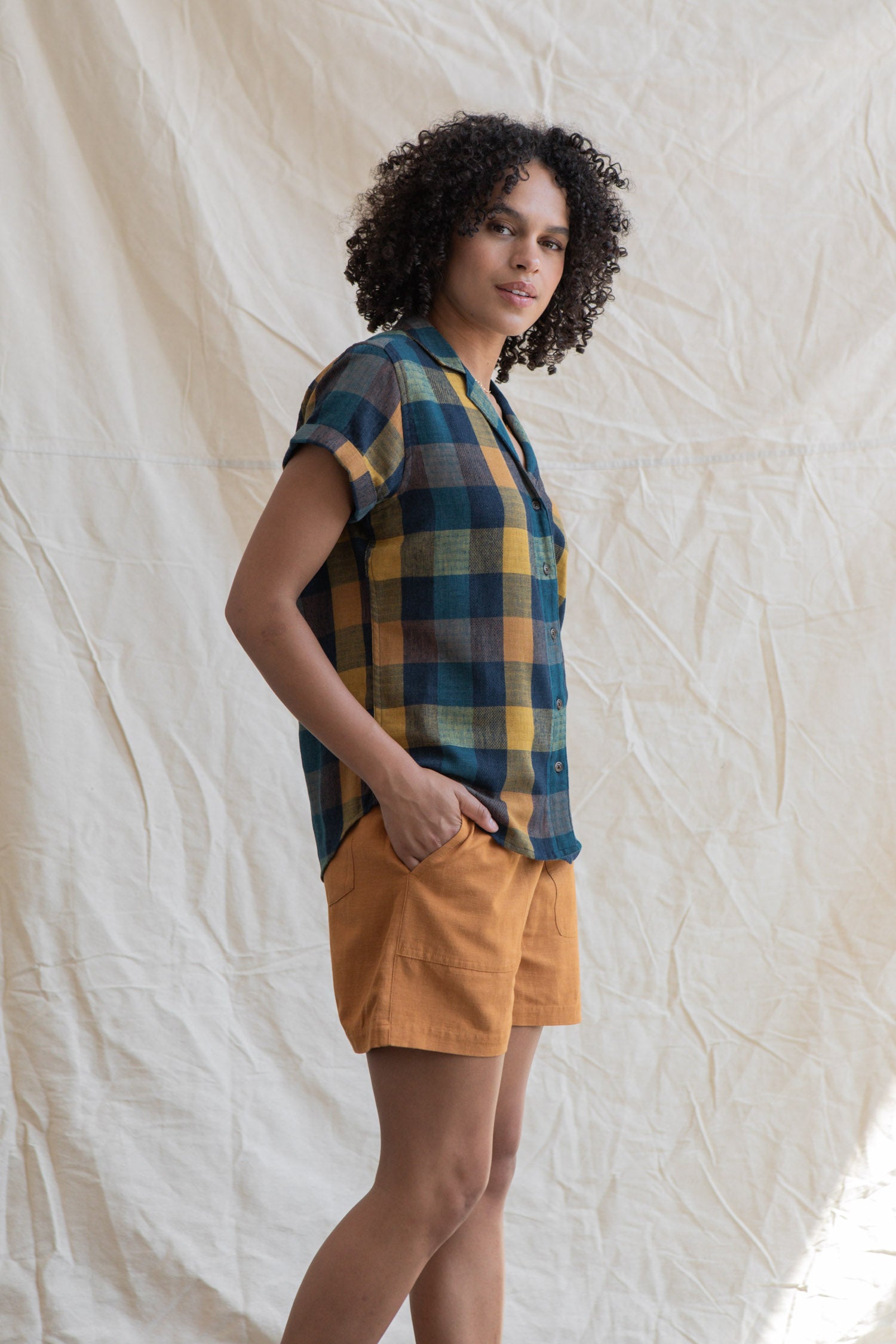A person with curly hair stands before a beige fabric backdrop, wearing the Bridge & Burn Innes Shirt in Moonbow Plaid— a relaxed fit, short-sleeve top made from recycled cotton—paired with mustard shorts, one hand in their pocket.
