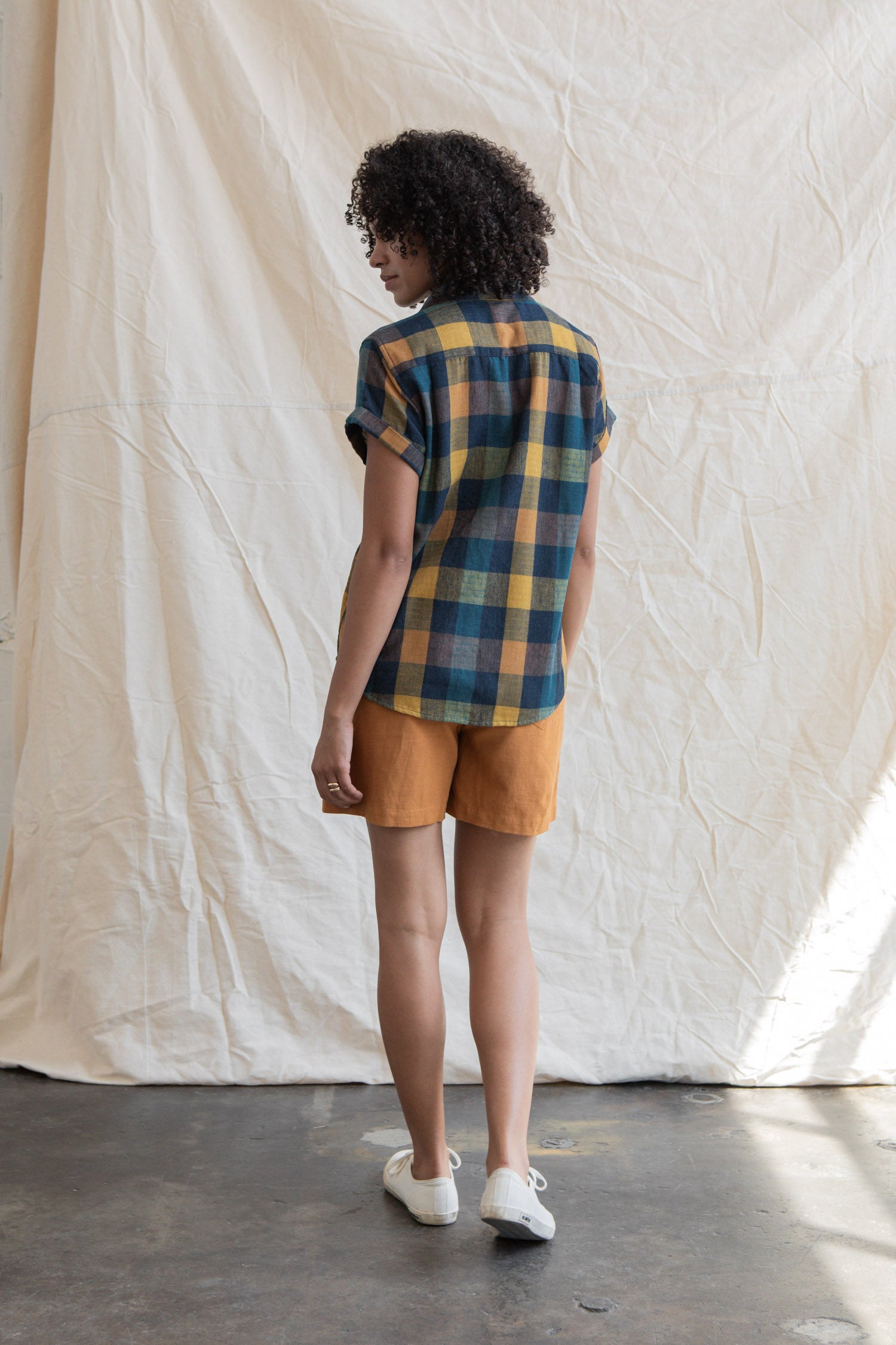 A person with curly hair, wearing the Bridge & Burn Innes Shirt in Moonbow Plaid and mustard shorts, stands indoors on a concrete floor facing away from the camera before a cream fabric backdrop.