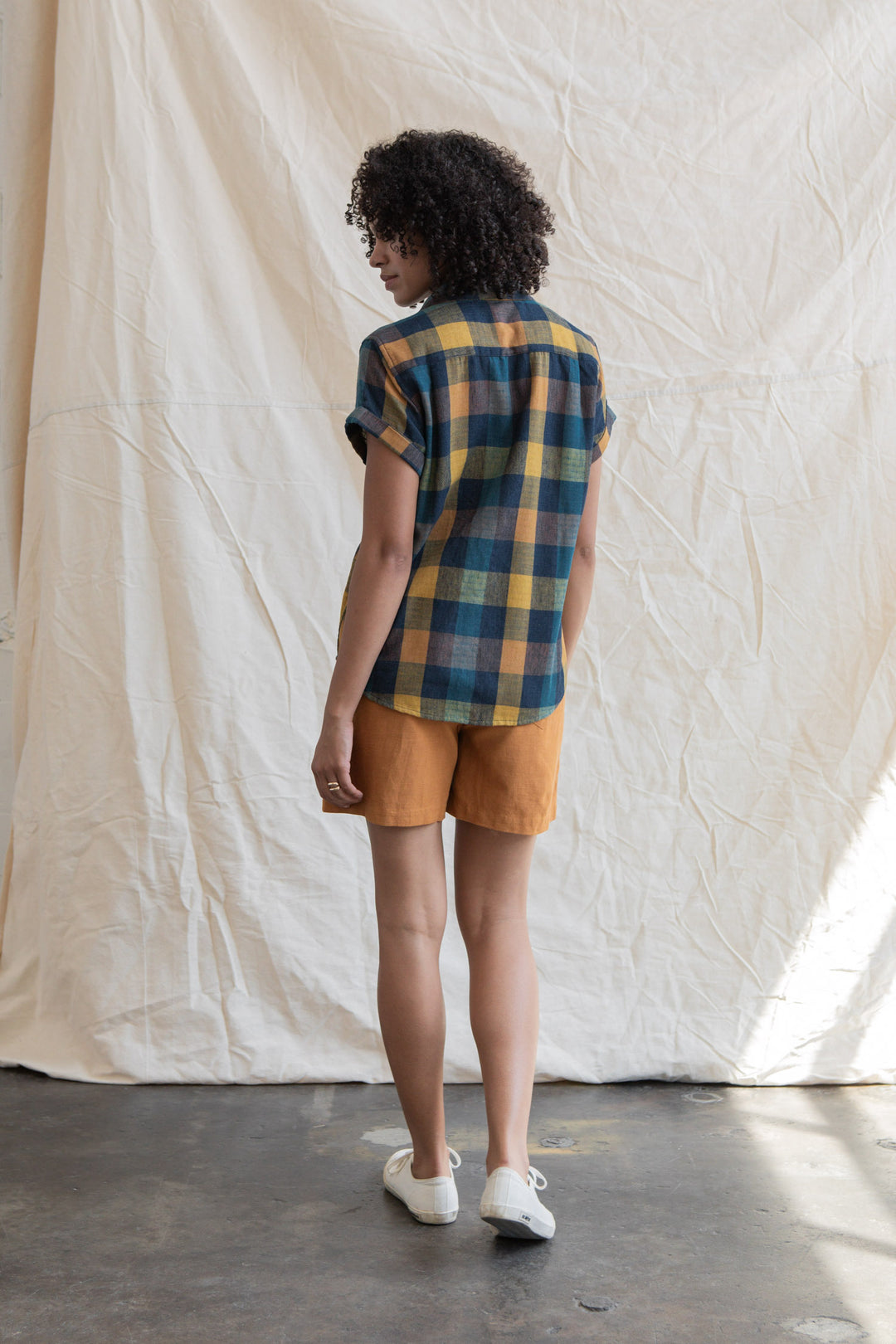 A person with curly hair, wearing the Bridge & Burn Innes Shirt in Moonbow Plaid and mustard shorts, stands indoors on a concrete floor facing away from the camera before a cream fabric backdrop.