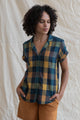 Wearing the Bridge & Burn Innes Shirt in Moonbow Plaid, a relaxed fit button-up of recycled cotton in blue, green, and yellow, a curly-haired person pairs it with mustard shorts, standing against a light beige fabric backdrop with hands in pockets.