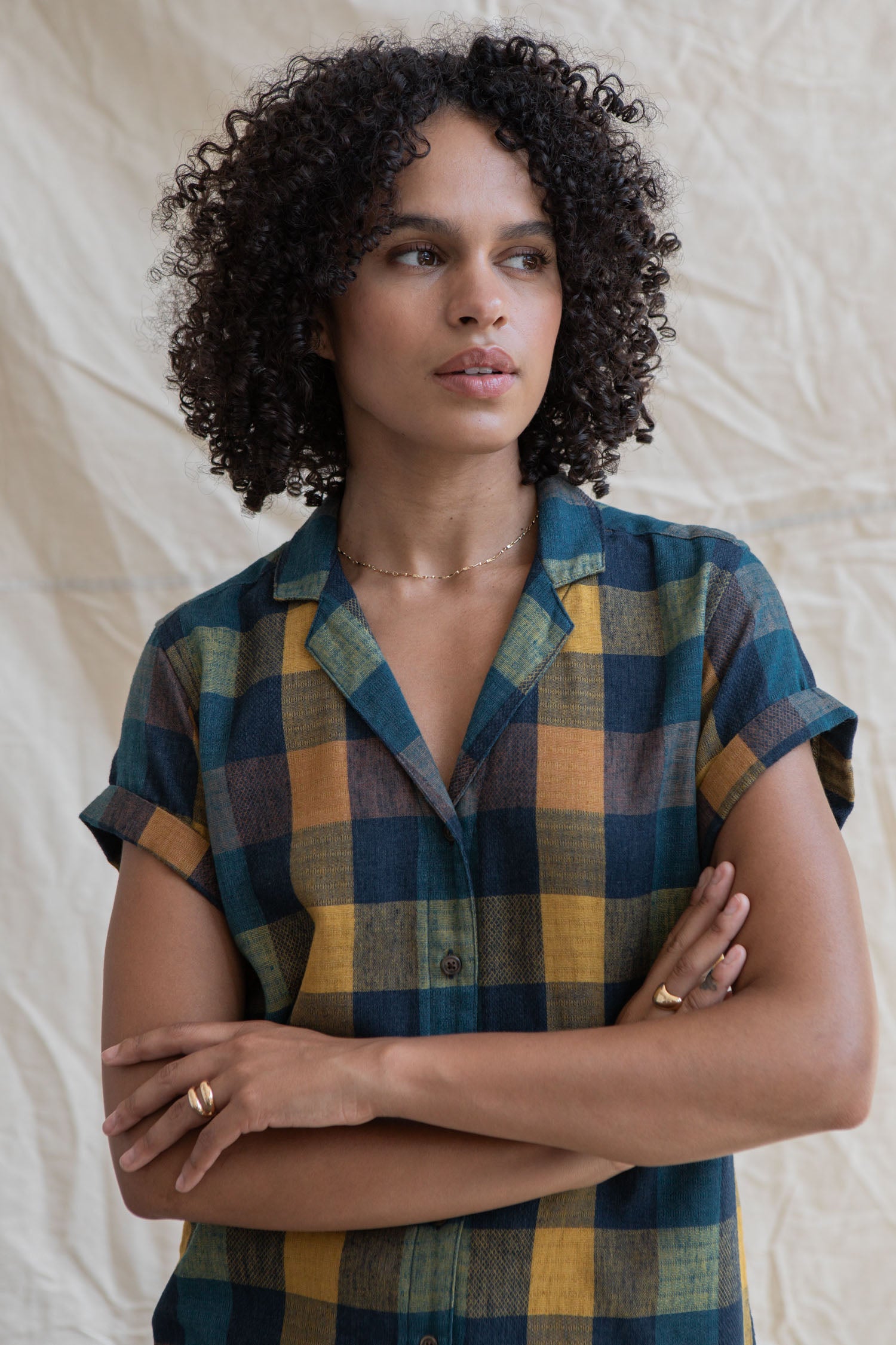 A person with curly hair stands with arms crossed, wearing the Bridge & Burn Innes Shirt in Moonbow Plaid against a neutral fabric background.