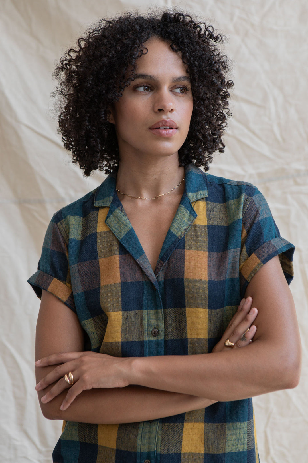 A person with curly hair stands with arms crossed, wearing the Bridge & Burn Innes Shirt in Moonbow Plaid against a neutral fabric background.