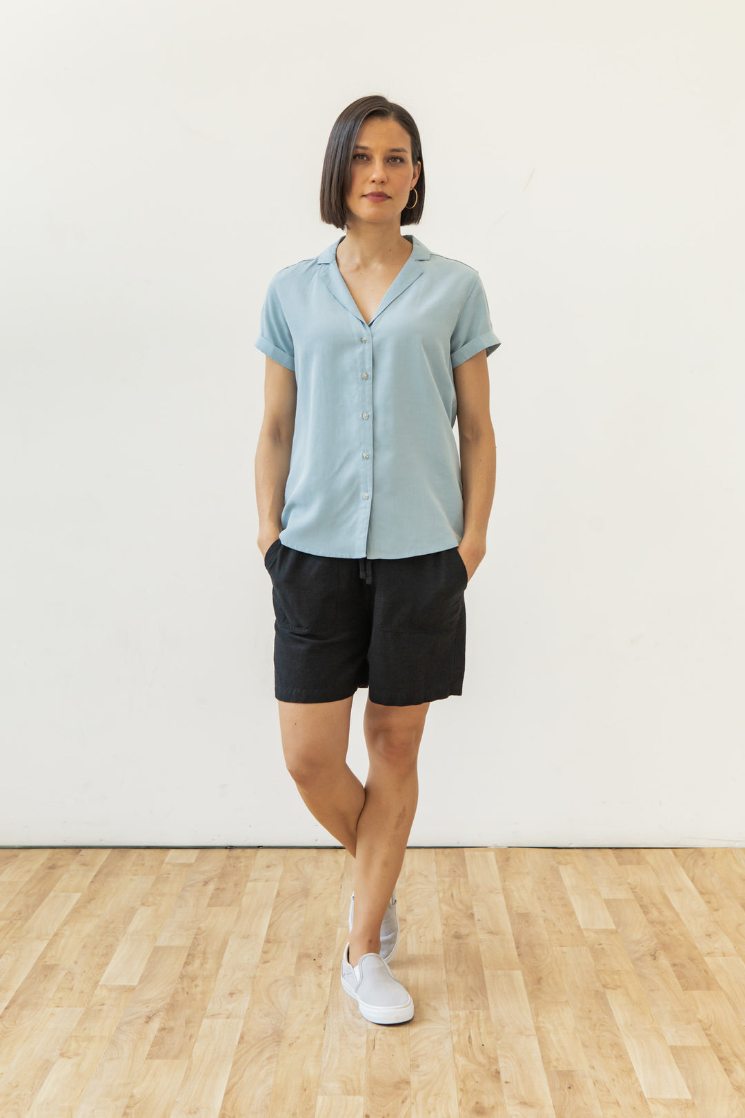 A woman stands against a plain white wall, wearing the Bridge & Burn Innes Shirt in Sky Blue with black shorts and white slip-ons. She crosses one leg over the other and keeps her hands in her pockets.