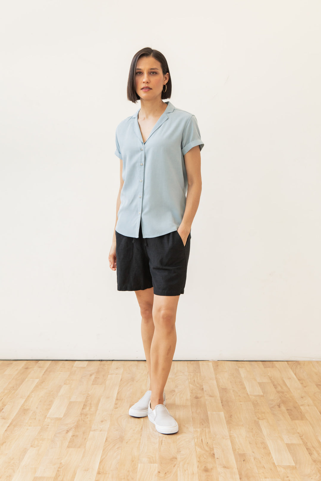 A woman with a short bob stands indoors against a white wall, wearing the Bridge & Burn Innes Shirt in Sky Blue, paired with black shorts and white slip-on shoes. She has one hand in her pocket and a neutral expression.