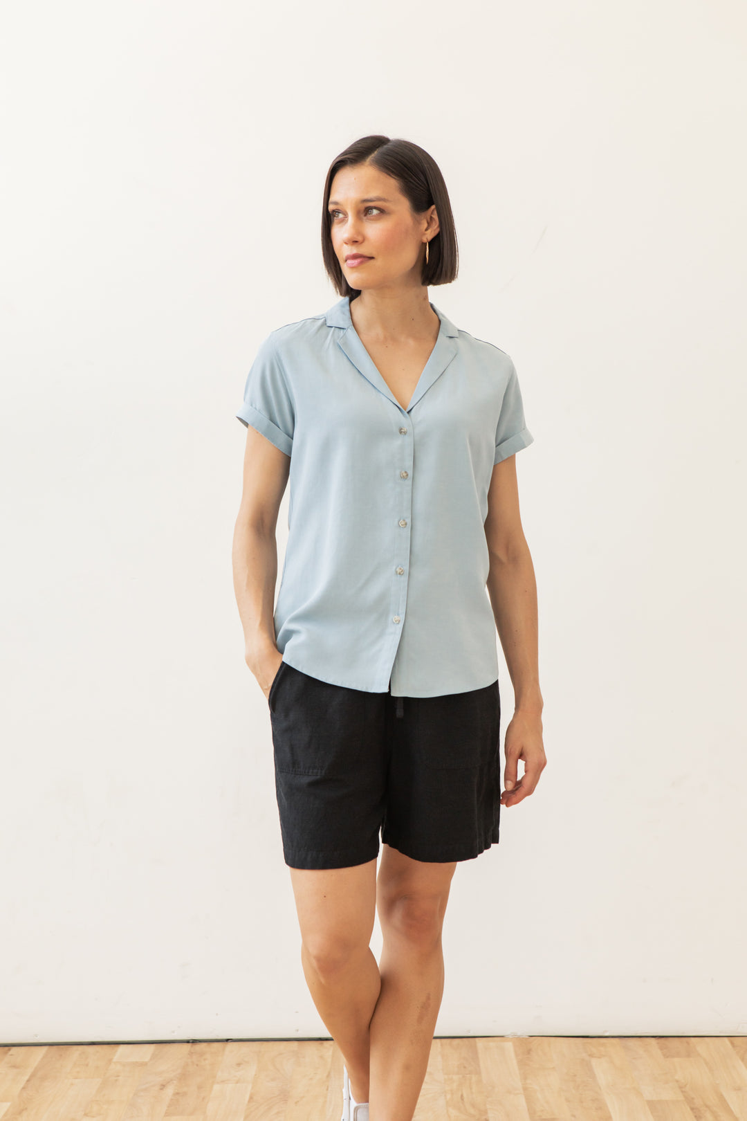A woman with short brown hair wears the Bridge & Burn Innes Shirt in Sky Blue and black shorts, standing on a wooden floor against a plain white background with one hand in her pocket.