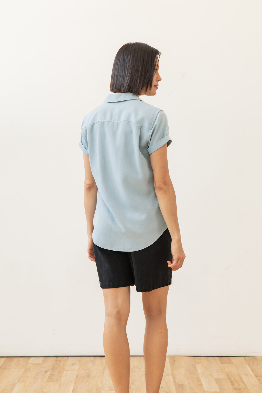 A woman with short dark hair faces away indoors, wearing the Bridge & Burn Innes Shirt in Sky Blue and black shorts. She stands on a wooden floor against a plain white wall.