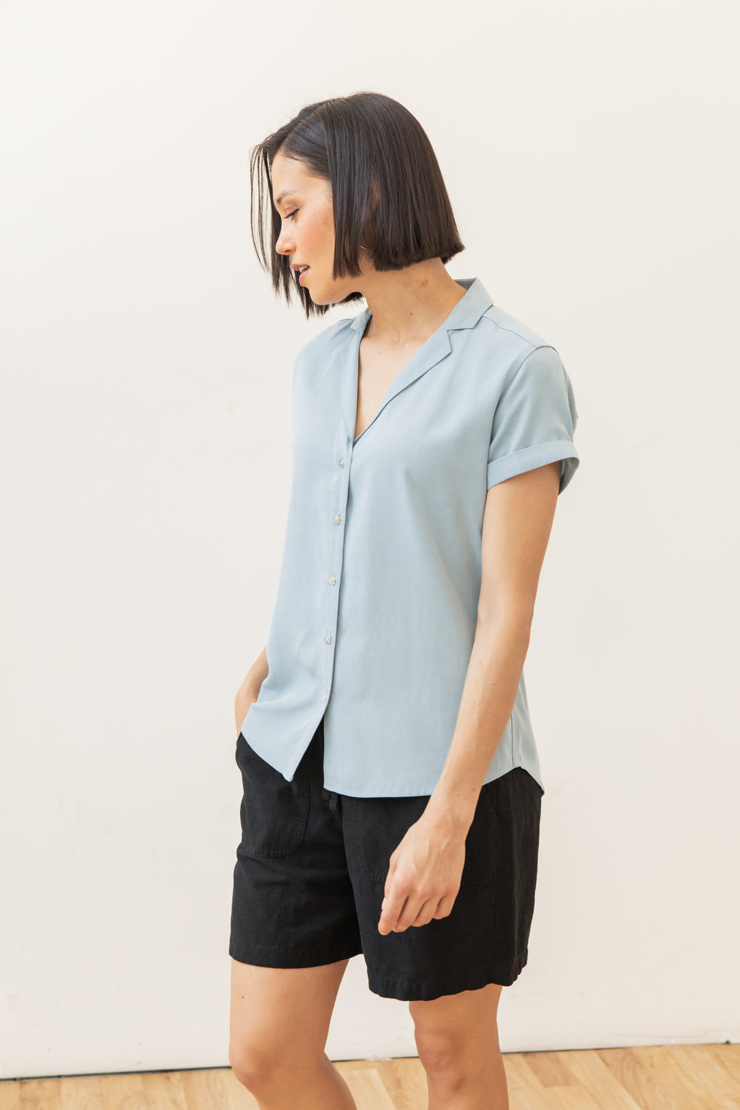 A woman with short dark hair wears the Bridge & Burn Innes Shirt in Sky Blue and black shorts, standing against a plain light background and looking down to her left.