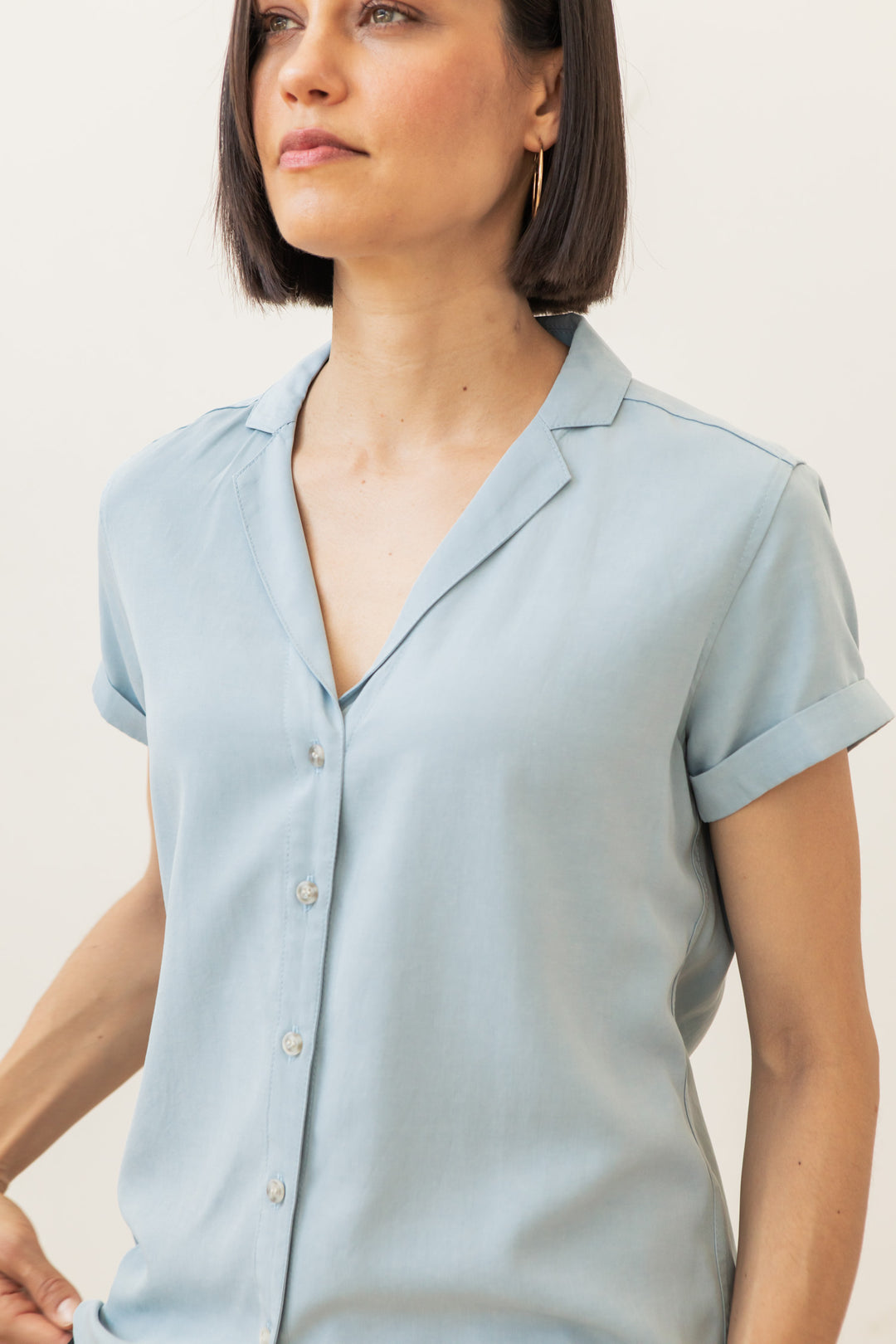 A woman with short dark hair wears the Bridge & Burn Innes Shirt in Sky Blue, a relaxed fit button-up, standing against a plain background and gazing slightly to the side.