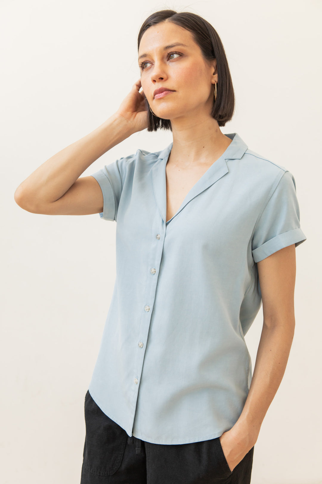 A woman with short brown hair wears the Bridge & Burn Innes Shirt in Sky Blue and black pants, standing against a plain background and touching her neck while looking slightly to the side.