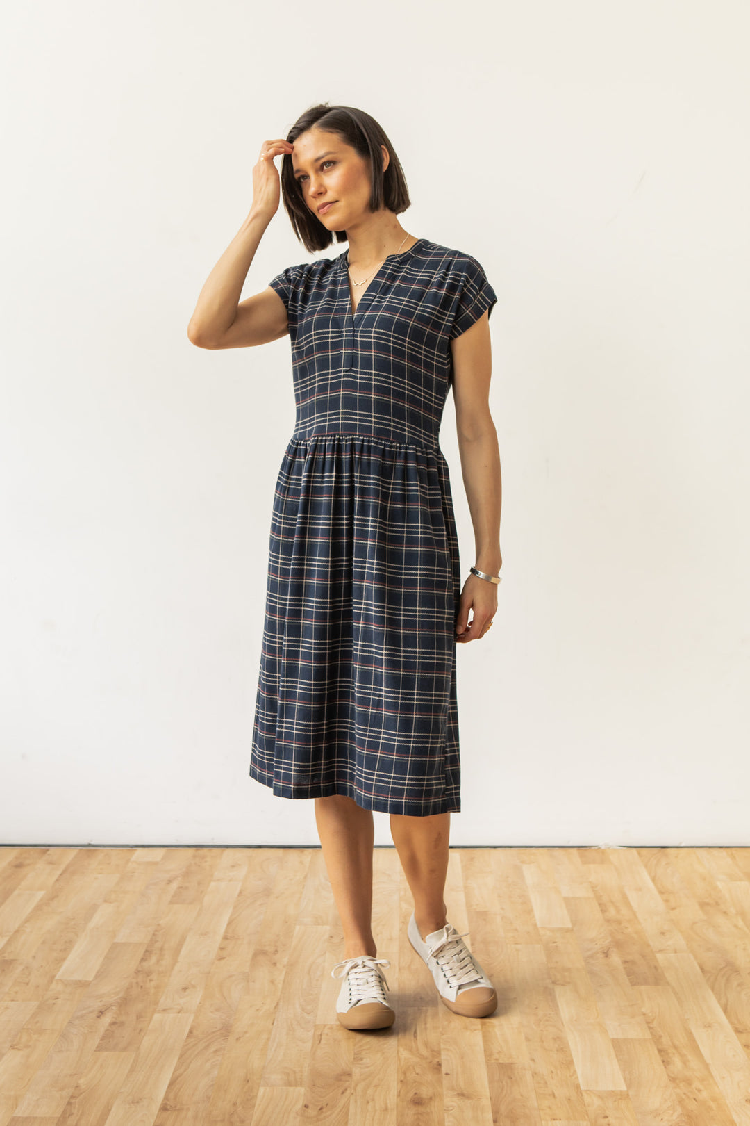 A woman wears the Bridge & Burn Ira Dress in Dusk Herringbone Plaid, a sustainable pullover made from recycled cotton, paired with tan sneakers. She stands on a wooden floor against a white wall, one hand touching her hair.