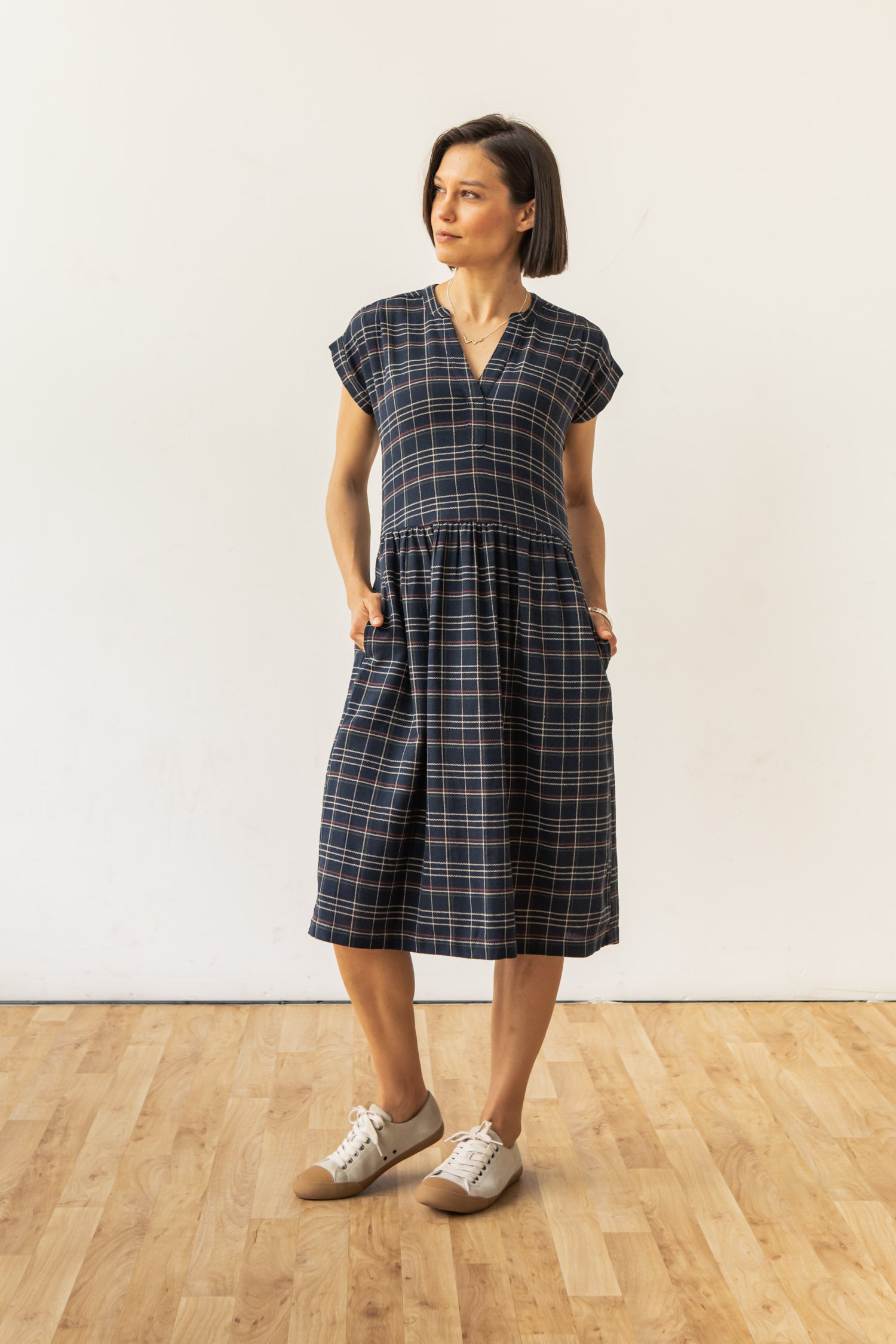 A woman stands indoors on a wooden floor, wearing the Bridge & Burn Ira Dress in Dusk Herringbone Plaid, a recycled cotton, short-sleeved, knee-length pullover with pockets, paired with beige sneakers. She looks slightly to the side with hands in her pockets.