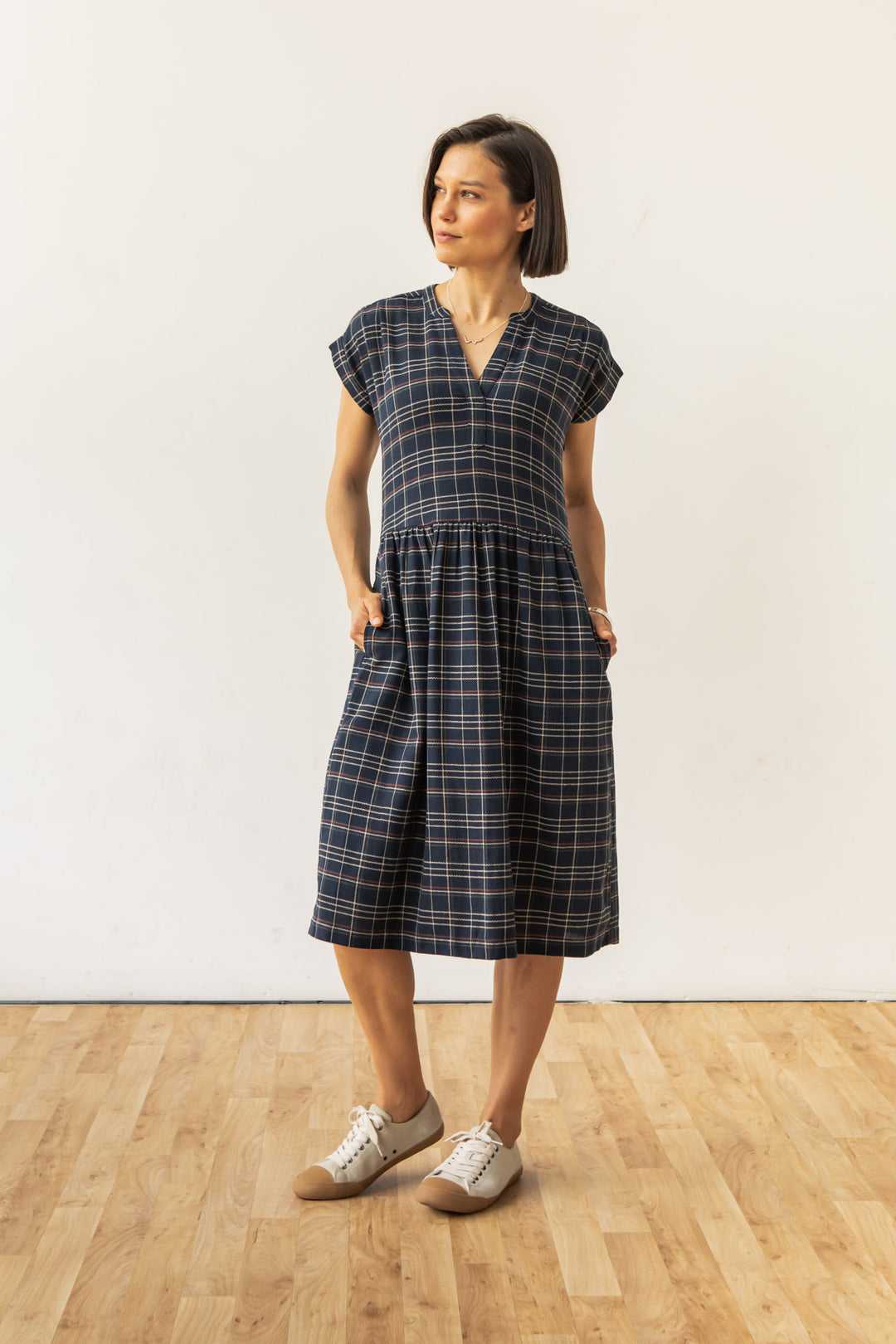 A woman stands indoors on a wooden floor, wearing the Bridge & Burn Ira Dress in Dusk Herringbone Plaid, a recycled cotton, short-sleeved, knee-length pullover with pockets, paired with beige sneakers. She looks slightly to the side with hands in her pockets.