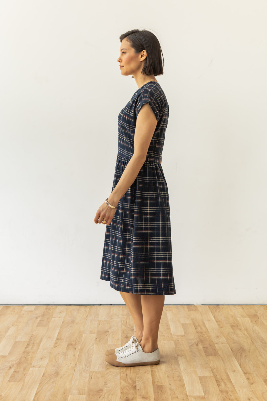 A woman stands in profile on a wooden floor by a white wall, wearing Bridge & Burn’s Ira Dress in Dusk Herringbone Plaid—crafted from recycled cotton—and white sneakers.