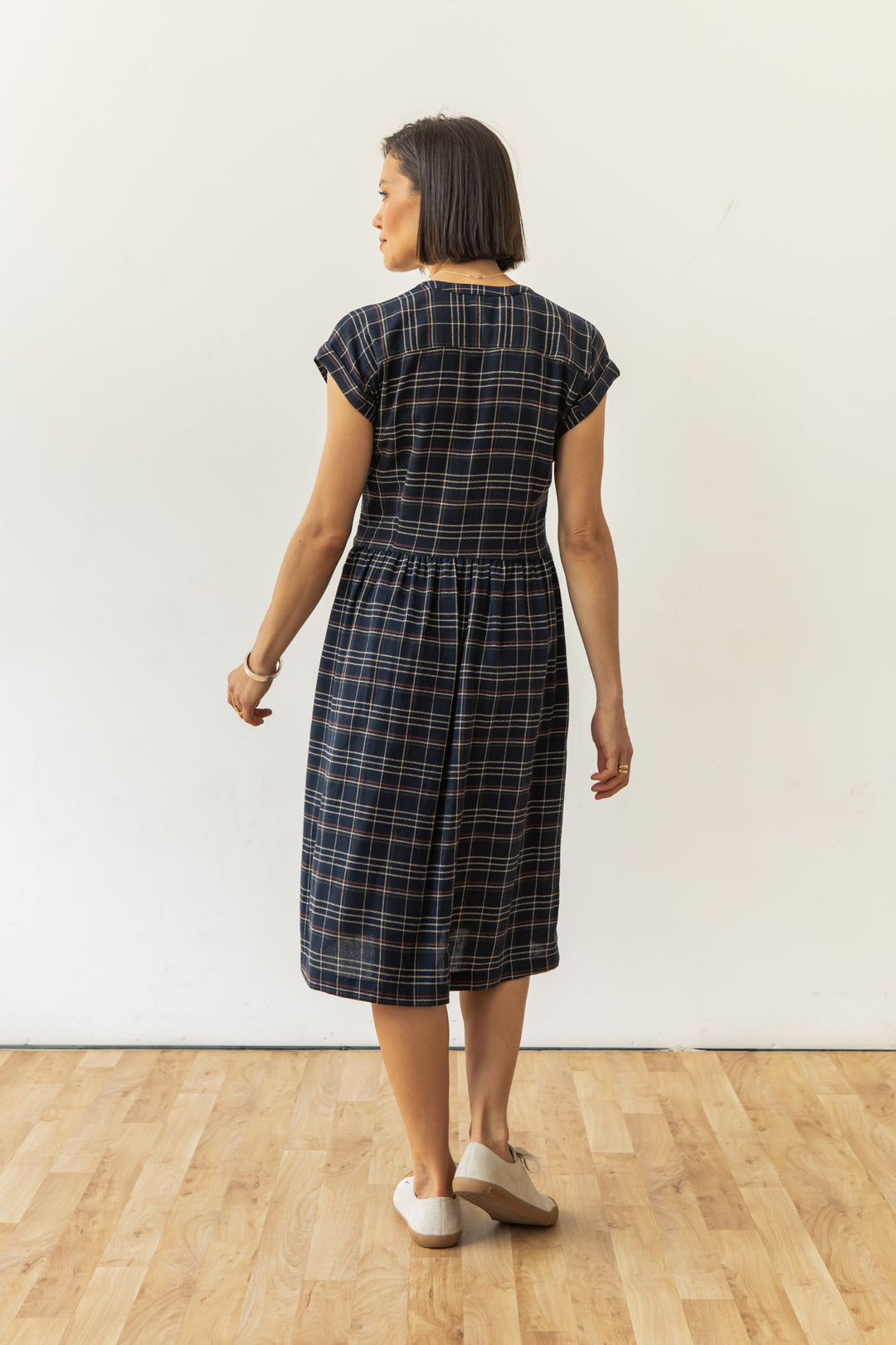A woman stands on a wooden floor against a white wall, wearing the Bridge & Burn Ira Dress in Dusk Herringbone Plaid and white shoes, facing away from the camera.