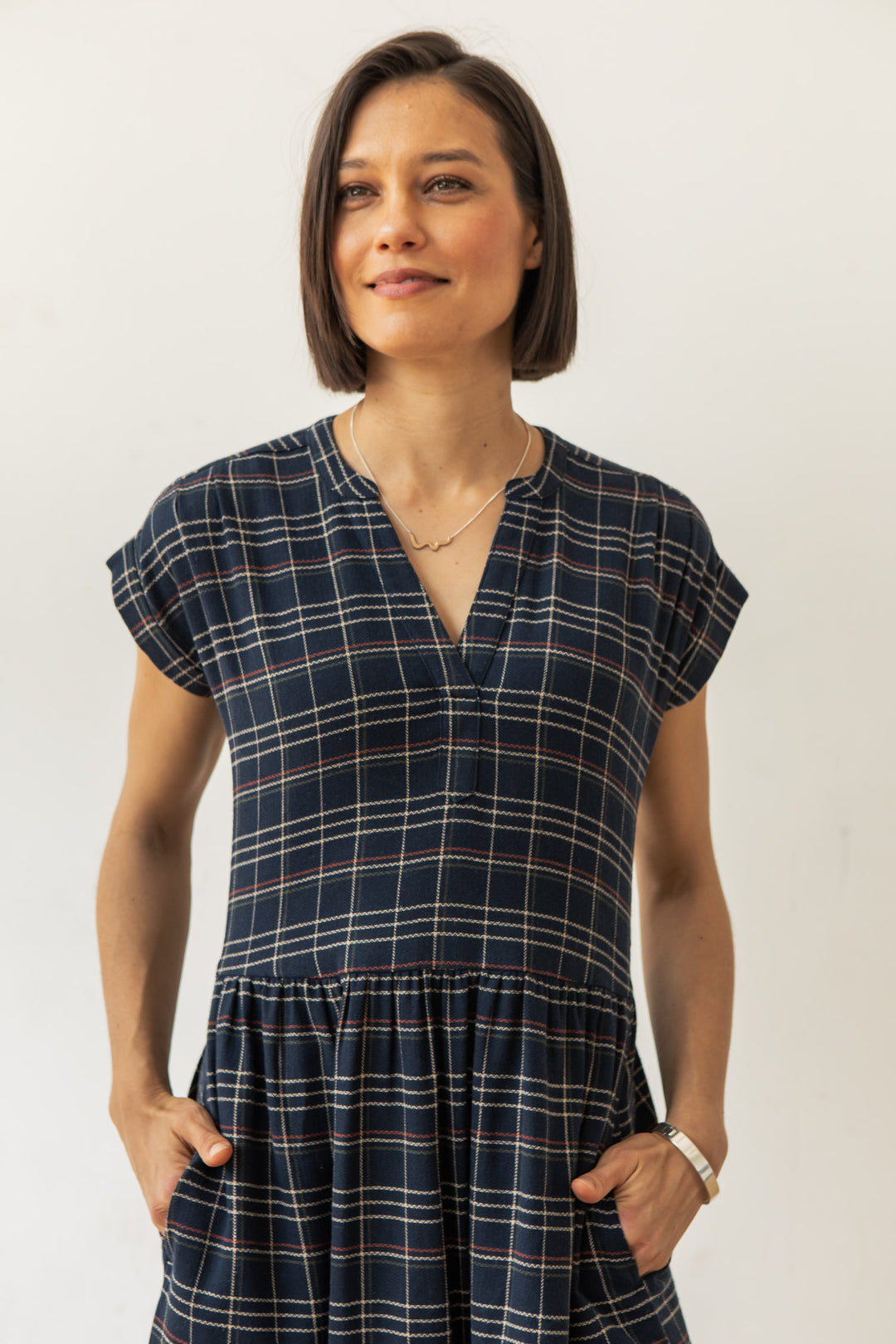 A woman with short brown hair stands against a plain light background, smiling slightly with her hands in the pockets of the Bridge & Burn Ira Dress in Dusk Herringbone Plaid.