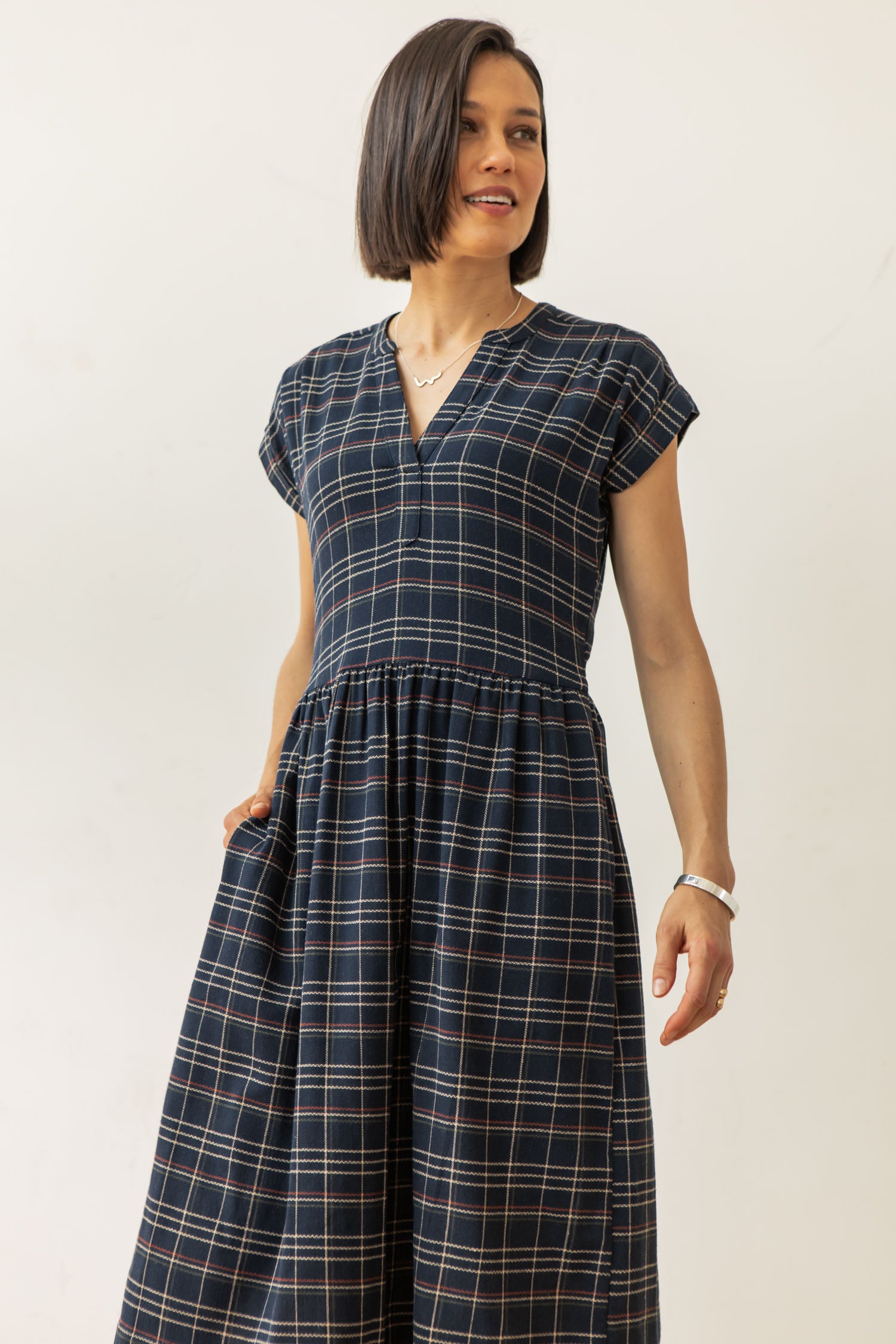 A woman with short dark hair models the Bridge & Burn Ira Dress in Dusk Herringbone Plaid, a navy V-neck pullover made from recycled cotton, standing against a plain light background with one hand in her pocket.
