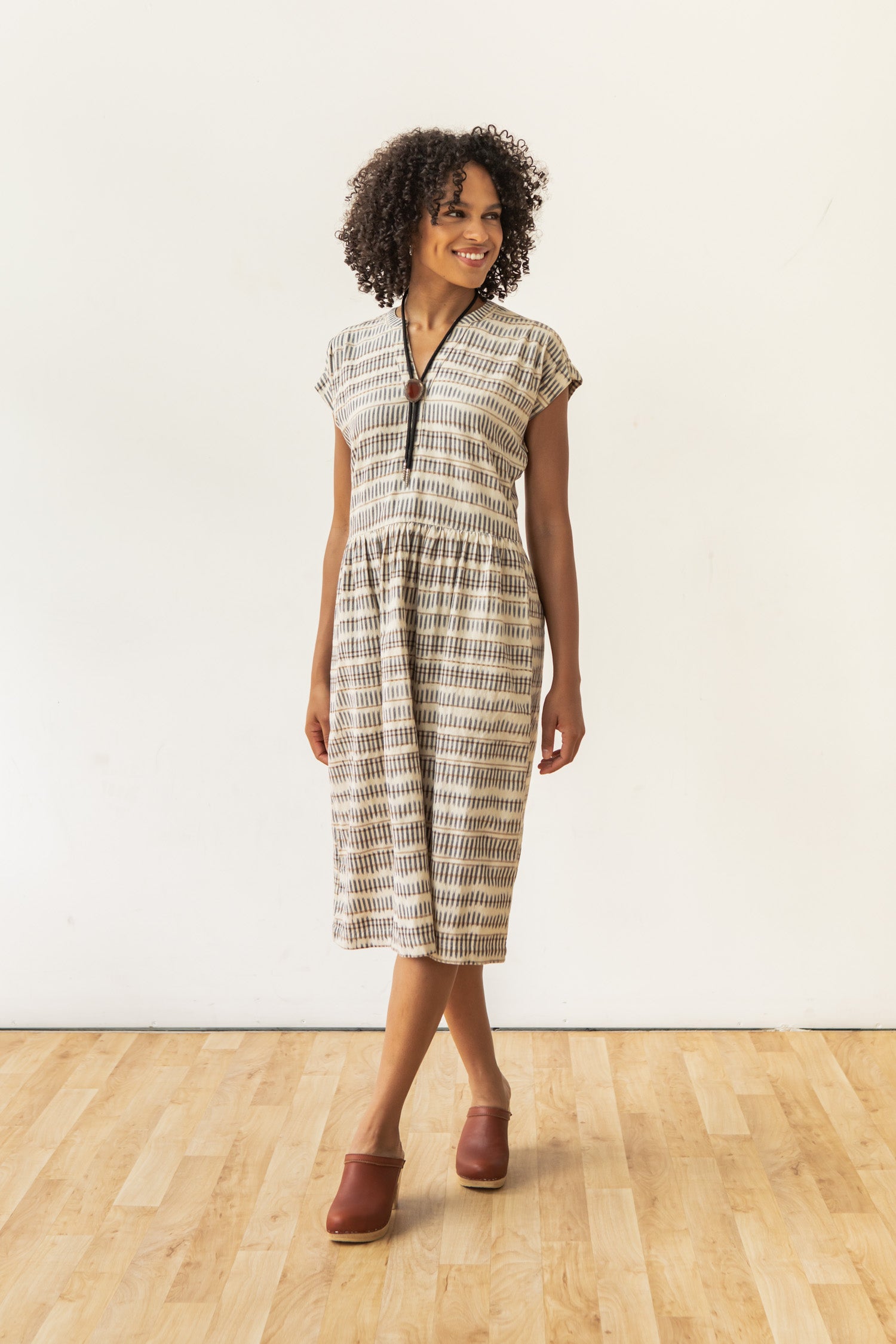 A woman with curly hair smiles on a wooden floor, wearing Bridge & Burn's Ira Dress in Mushroom Ikat Stripe—a patterned organic cotton, short-sleeve dress with a front zipper—paired with brown clogs against a plain white wall.