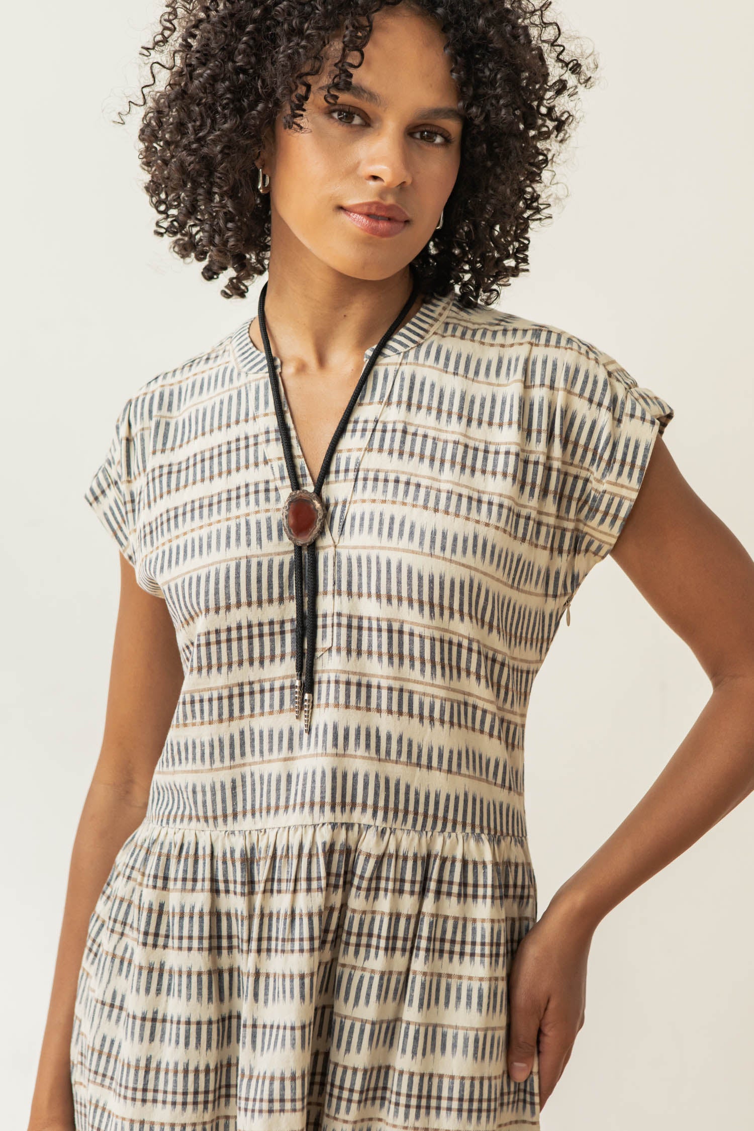 The Bridge & Burn Ira Dress in Mushroom Ikat Stripe is a short-sleeve, knee-length pullover with a V-neck, gathered waist, and neutral geometric stripes on lightweight fabric, shown laid flat on a white background.
