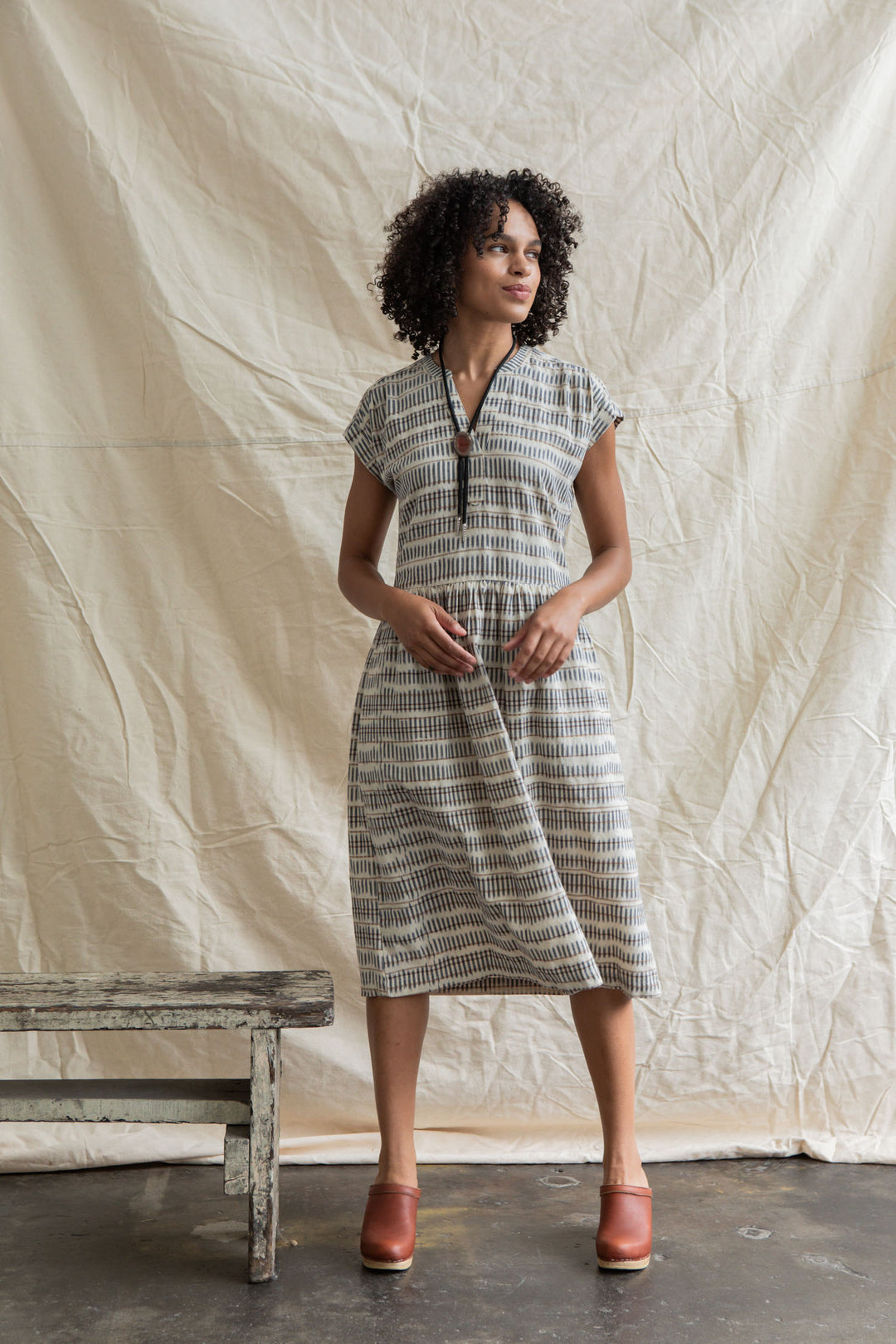 A woman with curly hair stands before a beige backdrop, wearing the Bridge & Burn Ira Dress in Mushroom Ikat Stripe and brown clogs. She poses with hands in her pockets near a rustic wooden bench.