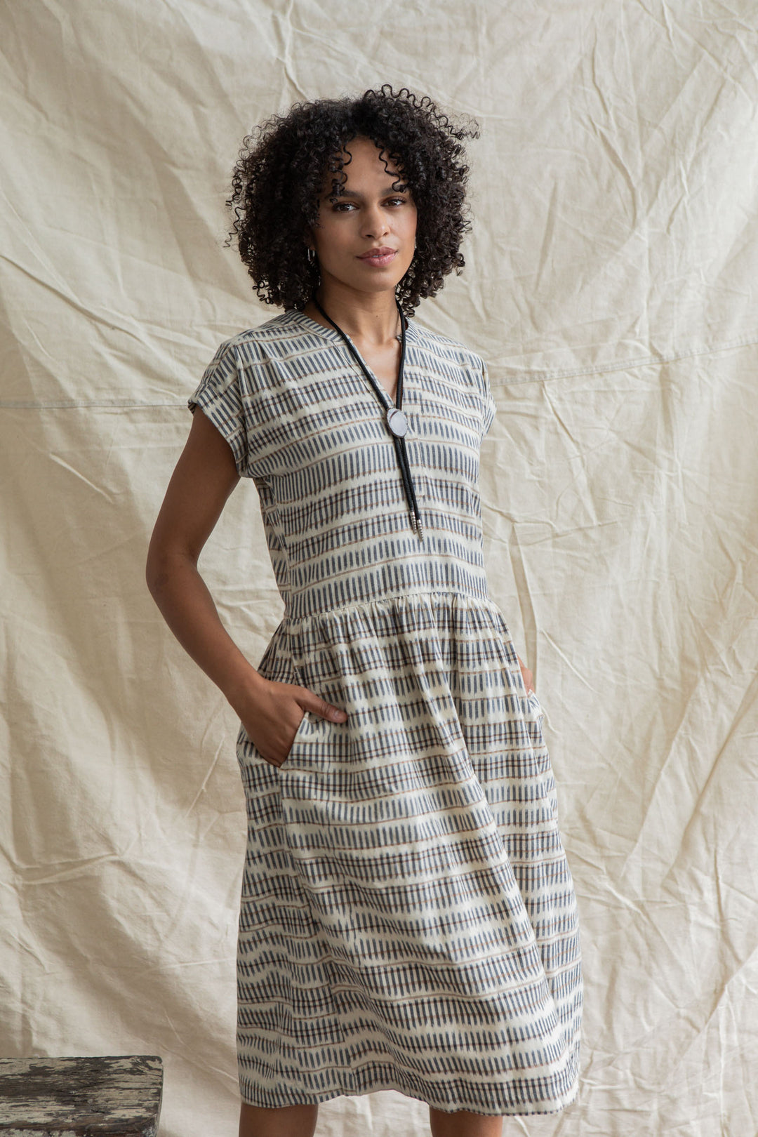 A woman with curly hair stands with her hands in the pockets of the Bridge & Burn Ira Dress in Mushroom Ikat Stripe, a knee-length, short-sleeved pullover with a gray and white geometric pattern, against a neutral textured backdrop.