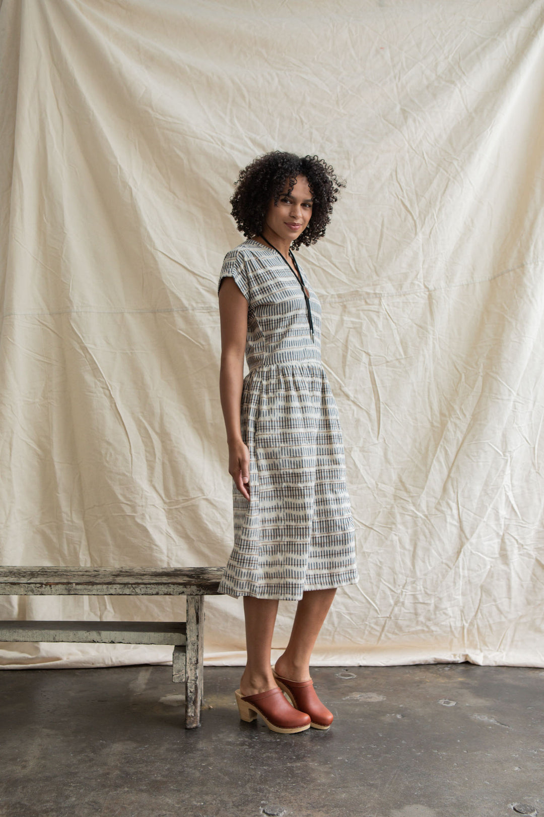 A woman with curly hair stands indoors before a cream fabric backdrop, wearing Bridge & Burn’s Ira Dress in Mushroom Ikat Stripe and brown clogs, resting one hand on a rustic wooden bench.