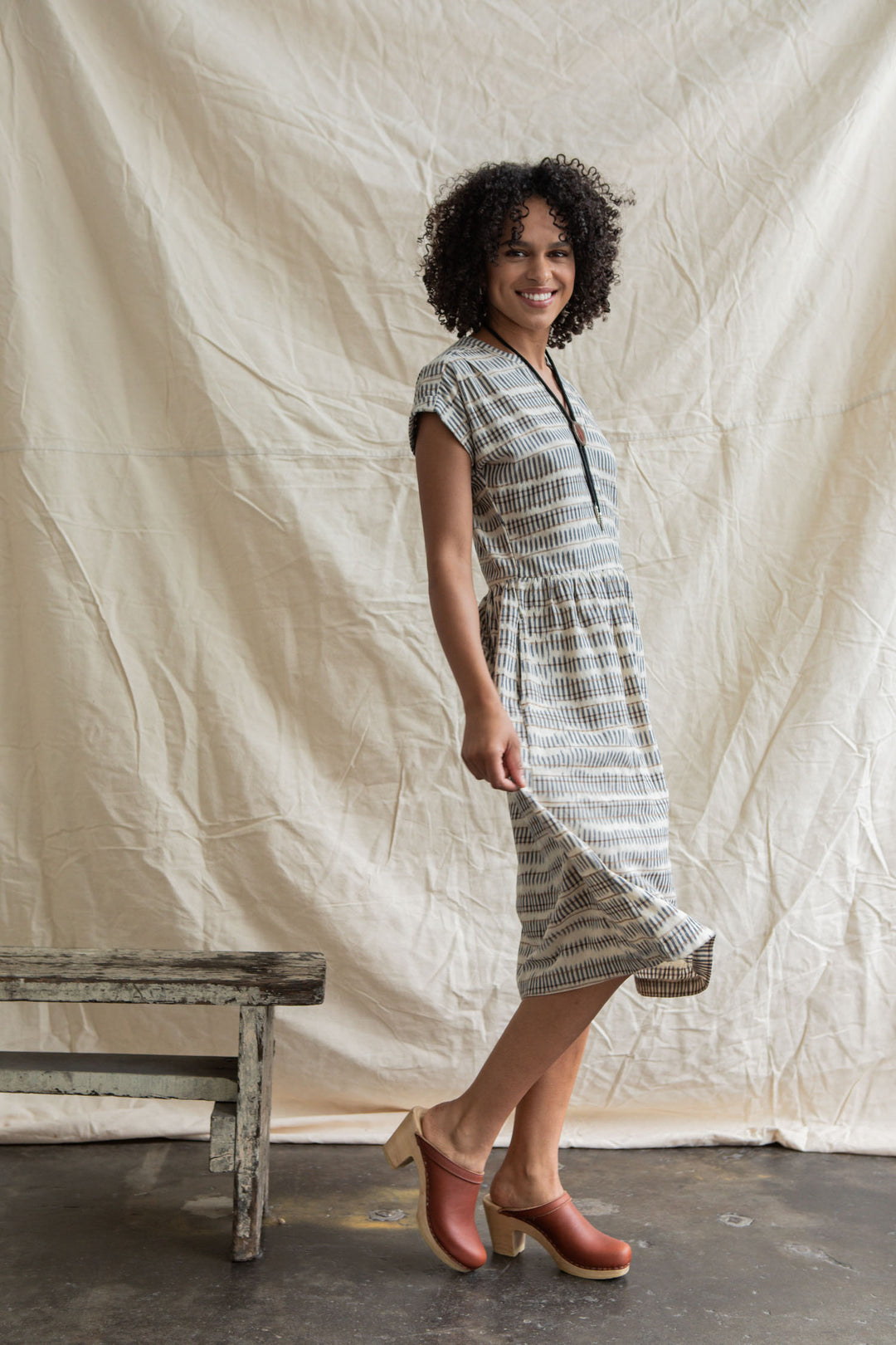 A woman with curly hair smiles, standing sideways and holding the hem of the Ira Dress in Mushroom Ikat Stripe by Bridge & Burn. She wears brown clogs, posing near a rustic bench against a wrinkled cream fabric backdrop.