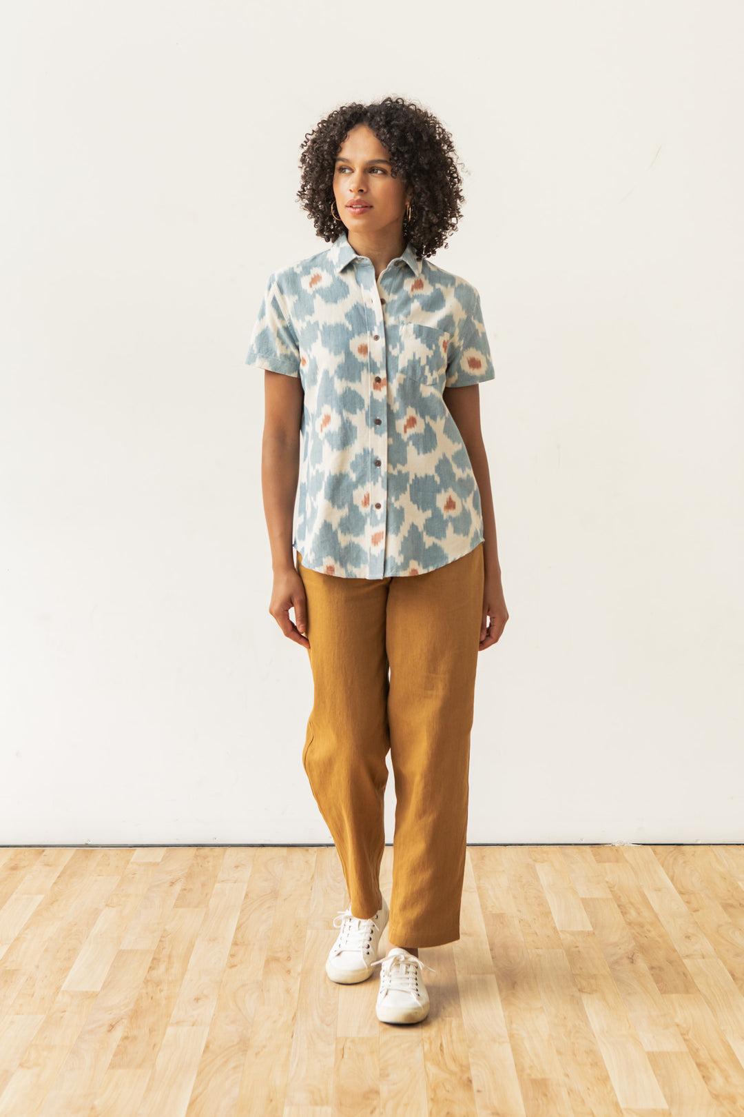 A woman with curly hair stands indoors on a wooden floor, wearing the Bridge & Burn Lana Shirt in Polar Blossom, paired with mustard-brown pants and white sneakers, looking slightly to the side.