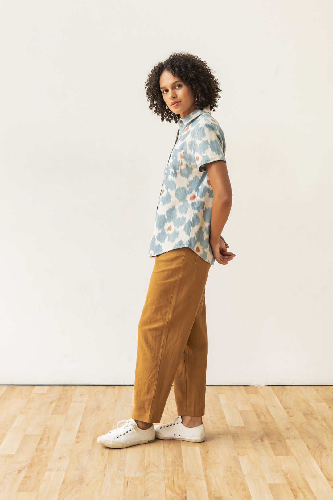 A person with curly hair stands sideways on a wooden floor wearing the Bridge & Burn Lana Shirt in Polar Blossom, mustard yellow pants, and white sneakers, against a plain white background.