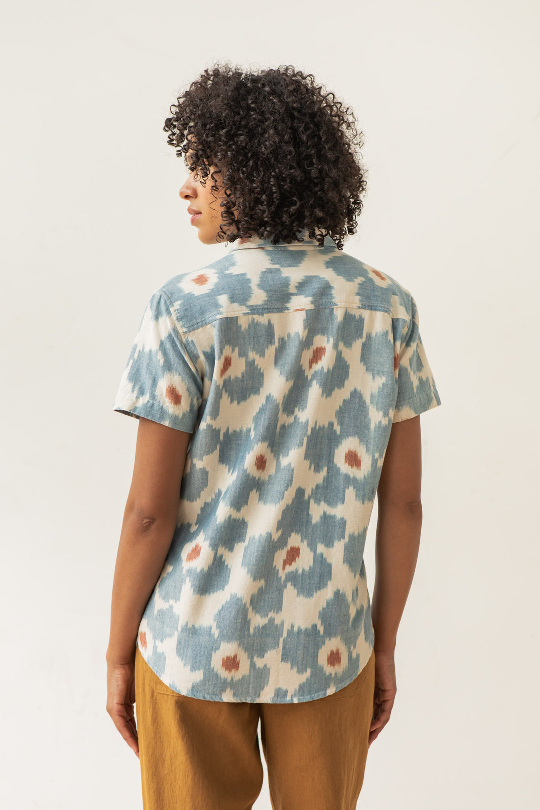 A person with short curly hair, seen from the back, wears the Bridge & Burn Lana Shirt in Polar Blossom—a relaxed fit, short sleeve button-up with a blue and beige abstract pattern—paired with brown pants against a plain light background.