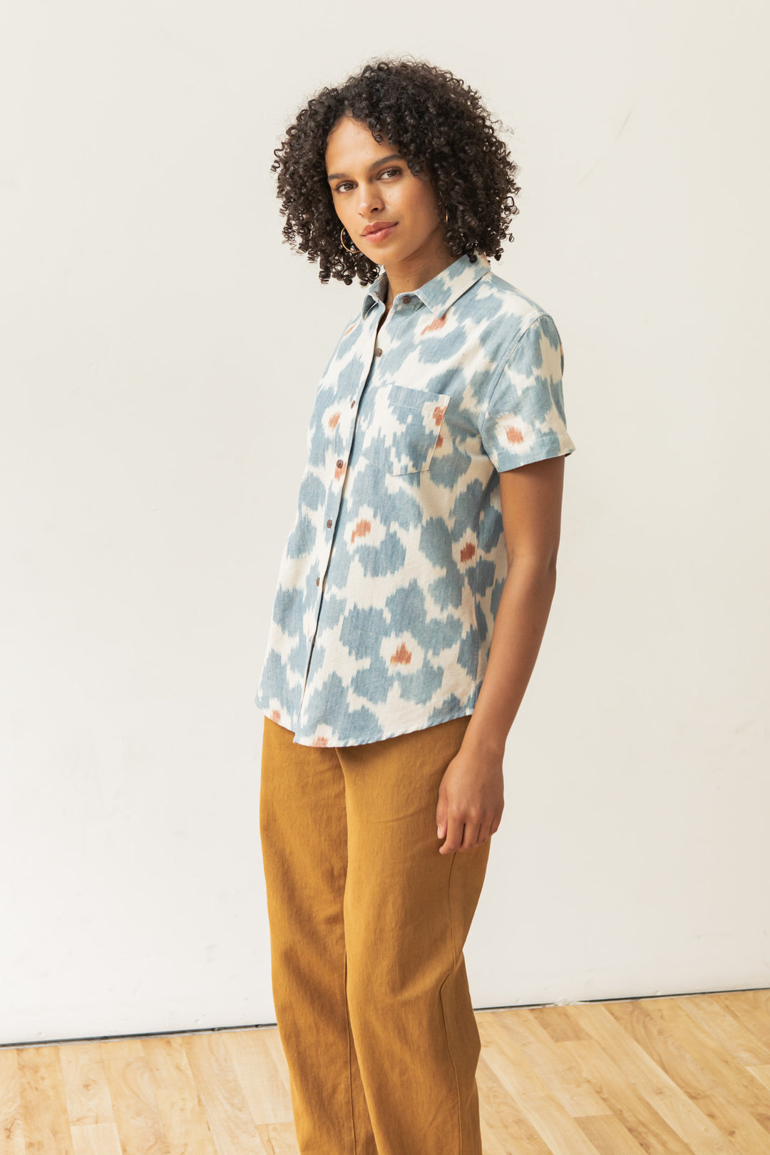 A person with curly hair stands indoors in the Bridge & Burn Lana Shirt / Polar Blossom, a relaxed fit short sleeve button up in blue and orange tones, paired with mustard yellow pants, against a plain light wall and wooden floor.