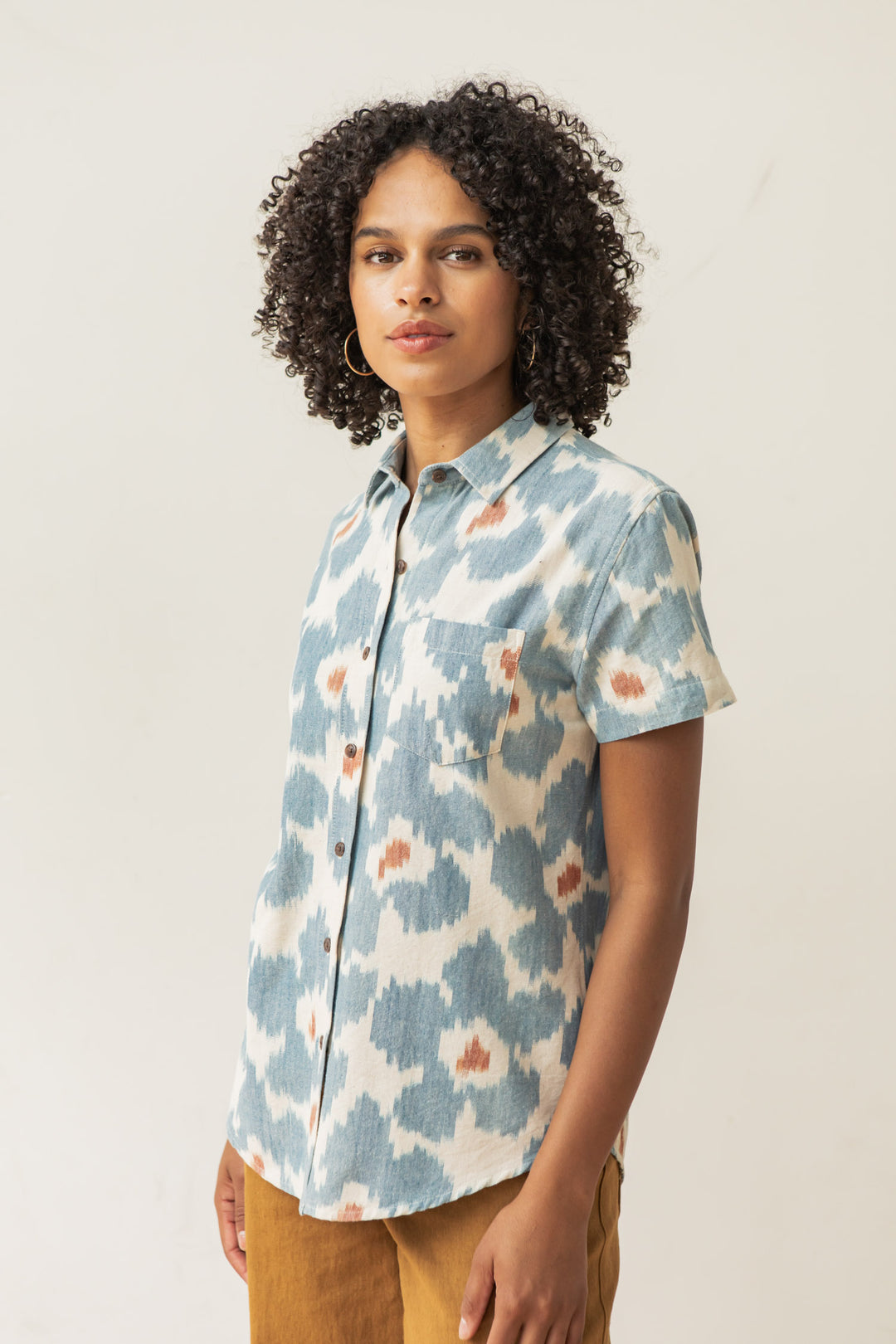 A person with curly hair wears the Bridge & Burn Lana Shirt in Polar Blossom—featuring a blue and white abstract pattern—and mustard pants, standing against a plain light background.