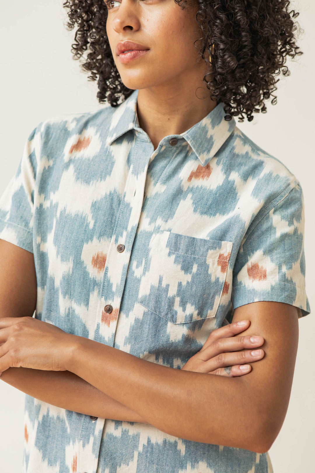 A person with curly hair wears the Bridge & Burn Lana Shirt in Polar Blossom, a relaxed fit, short sleeve 100% cotton button-up with a blue and white ikat pattern and brown accents, standing arms crossed against a light background.