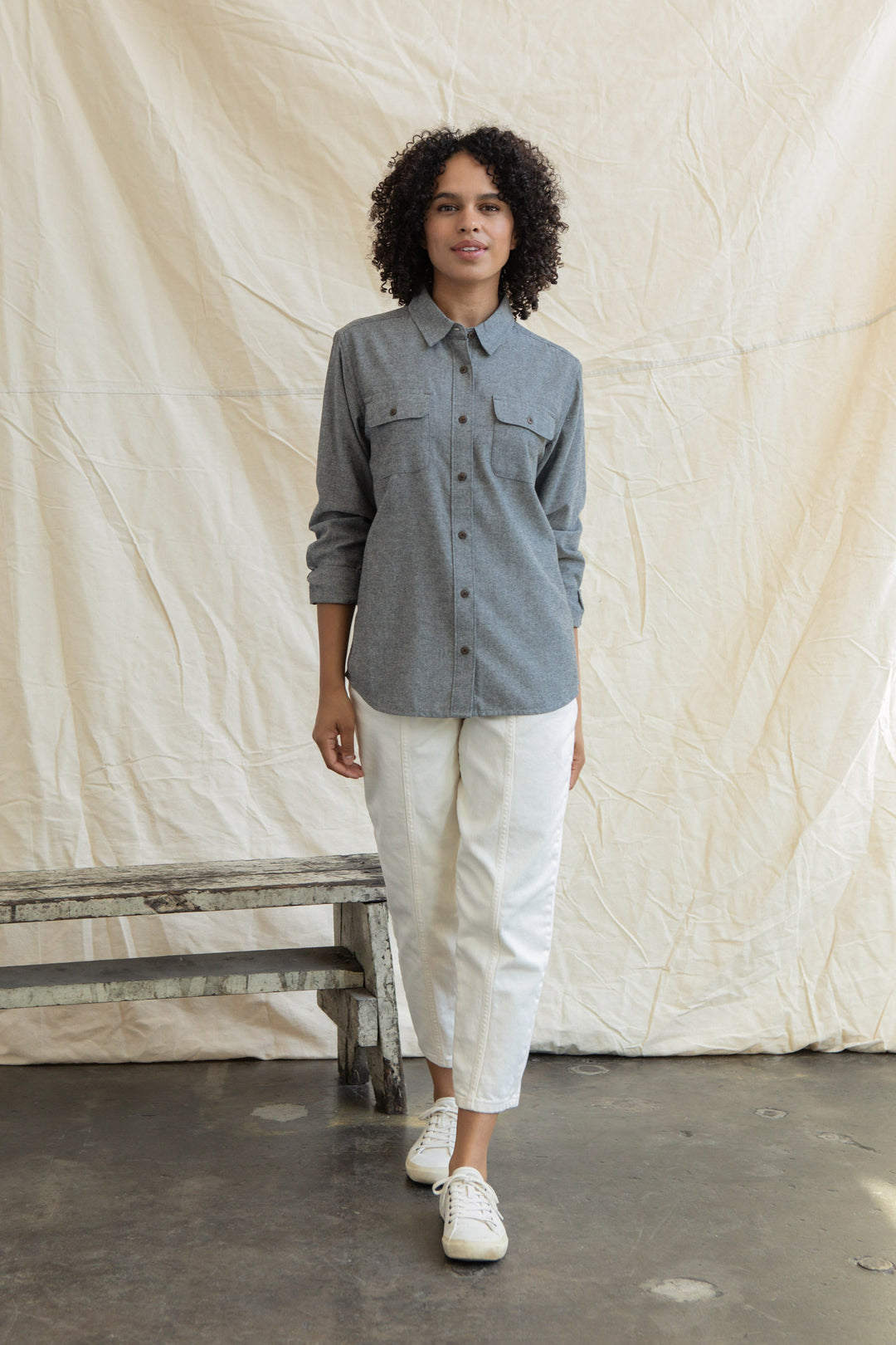 A person with curly hair wears the Bridge & Burn Lenon Shirt in Iron Chambray, paired with white pants and white sneakers, standing indoors on a concrete floor beside a rustic wooden bench and a cream-colored fabric backdrop.