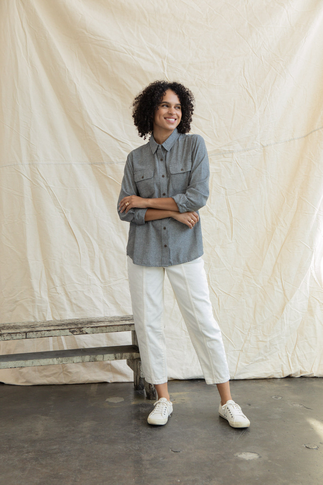 A person with curly hair smiles in front of a cream fabric backdrop, wearing the Bridge & Burn Lenon Shirt in Iron Chambray, paired with white pants and sneakers, arms loosely crossed.