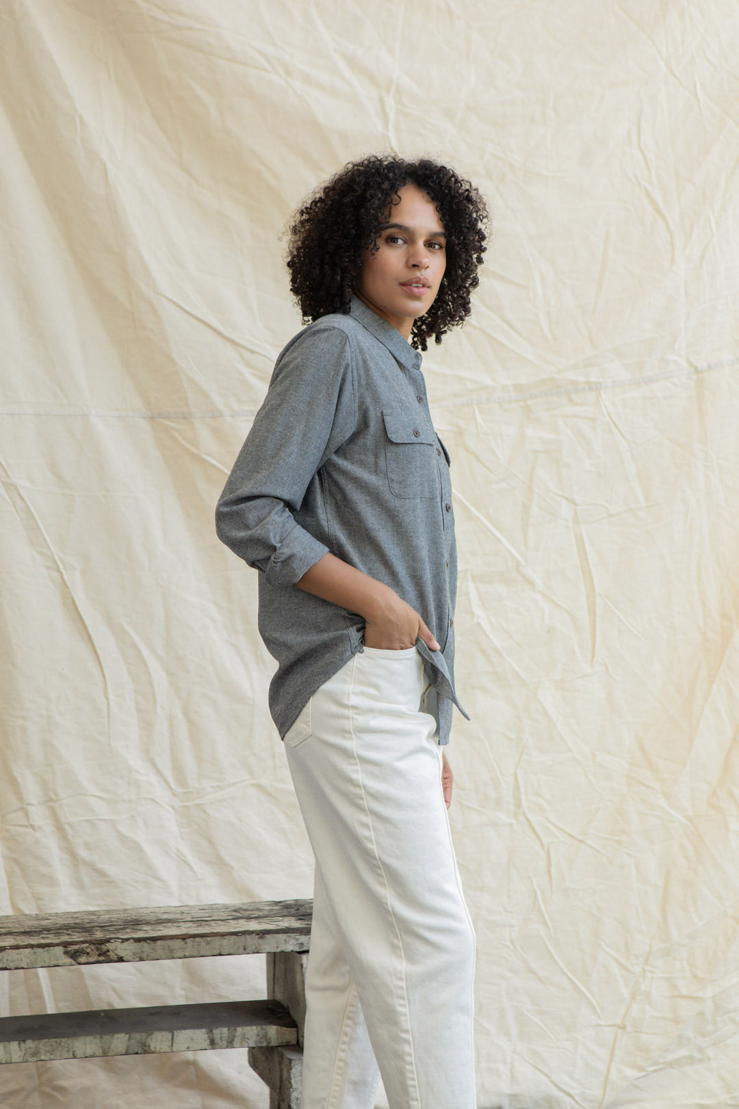 A person with curly hair stands before a beige fabric backdrop, wearing the Bridge & Burn Lenon Shirt in Iron Chambray and white pants, one hand in their pocket, looking confidently at the camera.